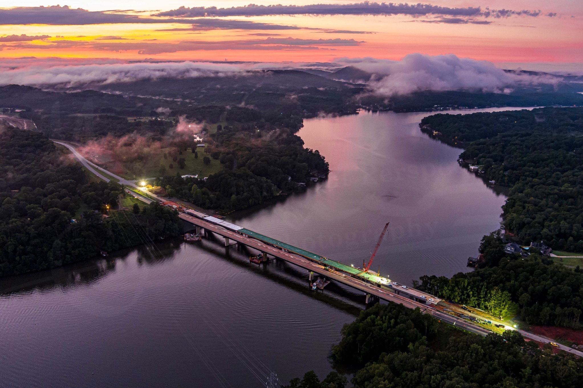 Aerial view of a bridge construction site over a river at sunset, surrounded by fog and lush trees