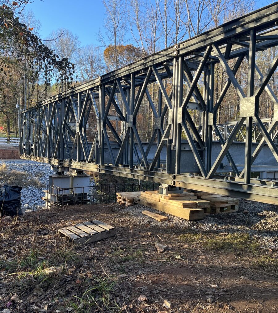 Metal truss bridge under construction in a wooded area, clear sky