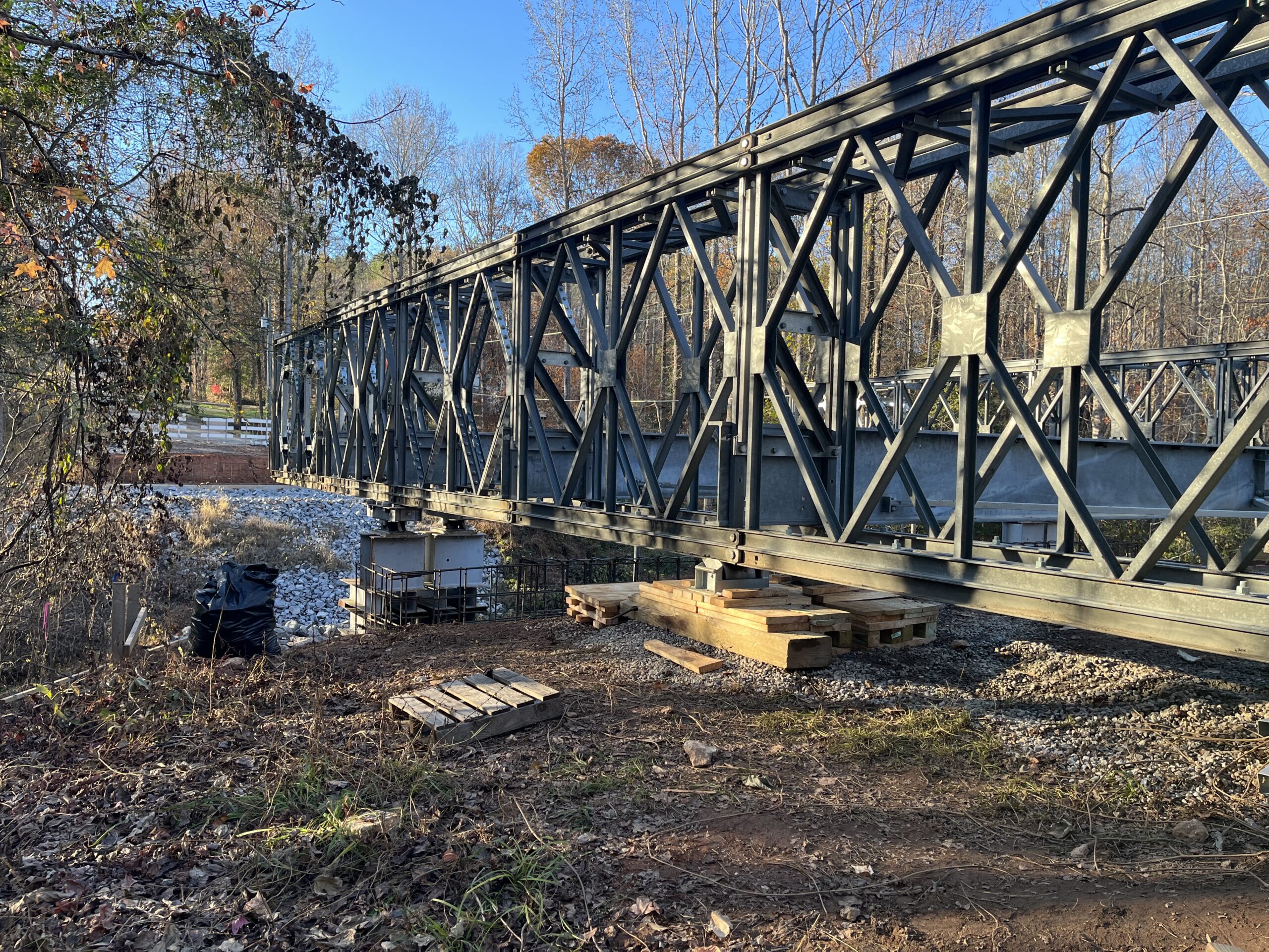 Metal truss bridge under construction in a wooded area, clear sky