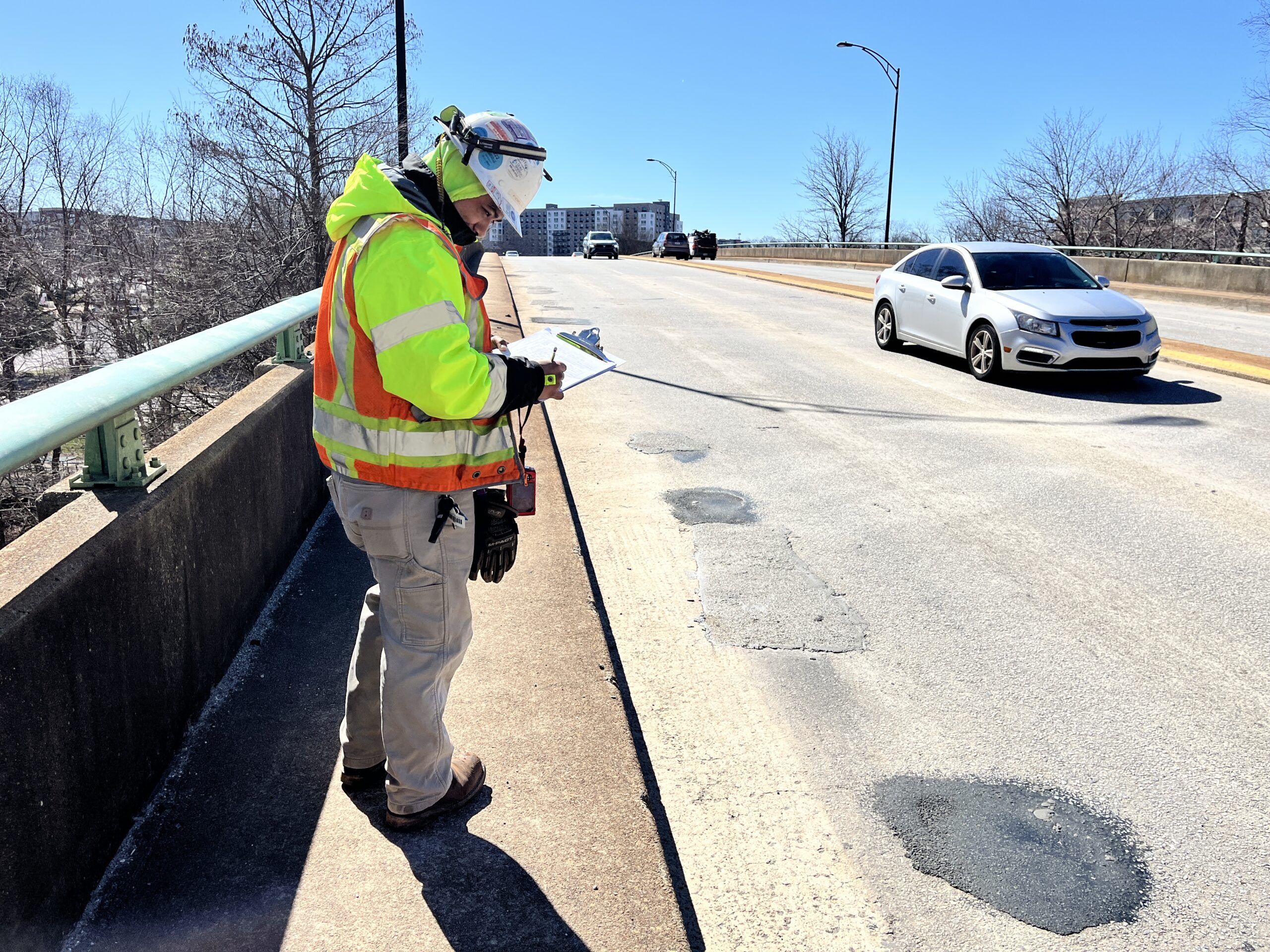 Construction worker inspecting road potholes on a bridge, writing notes on clipboard