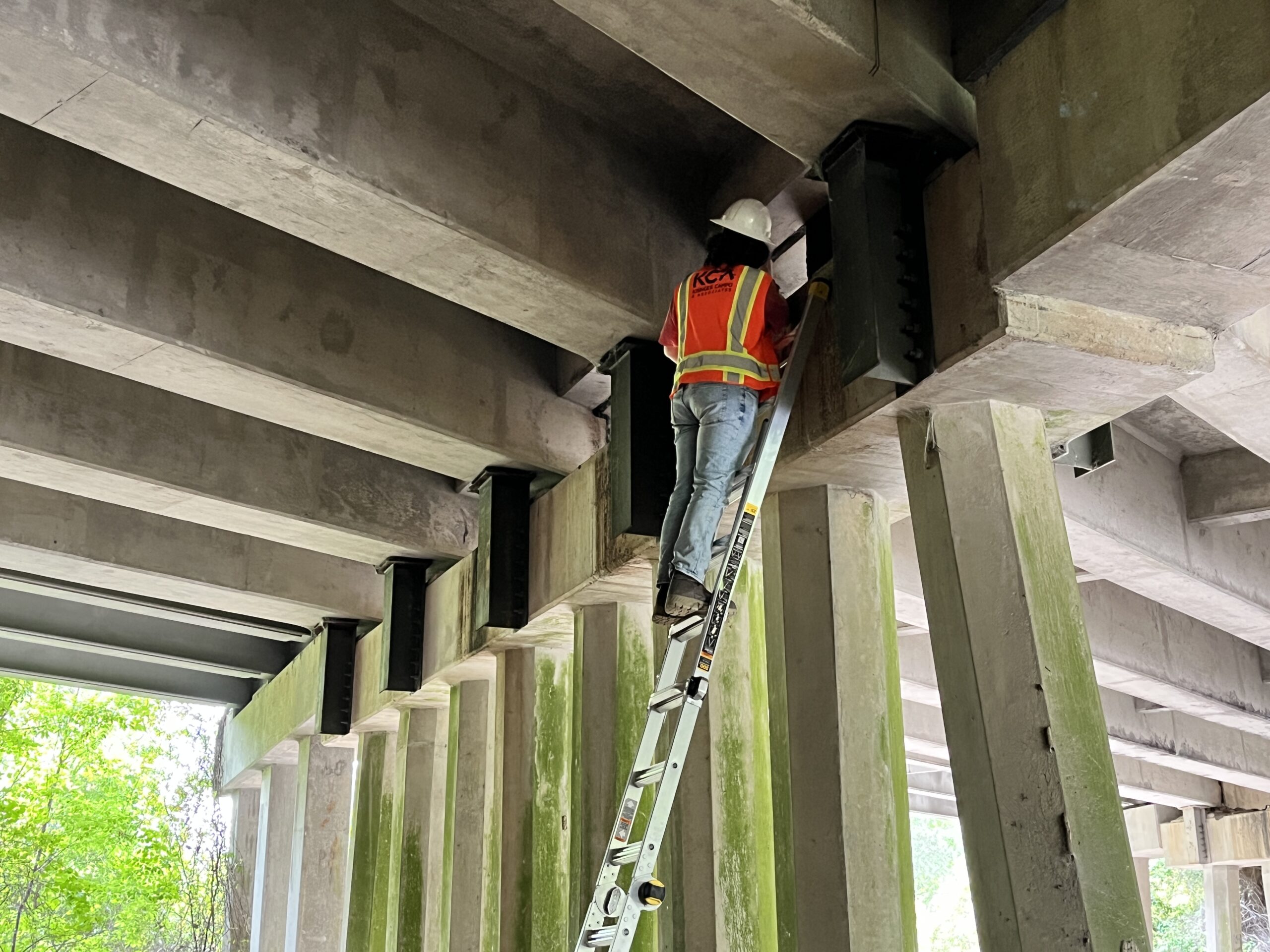Person in a safety vest and hard hat inspecting bridge underside, standing on a ladder