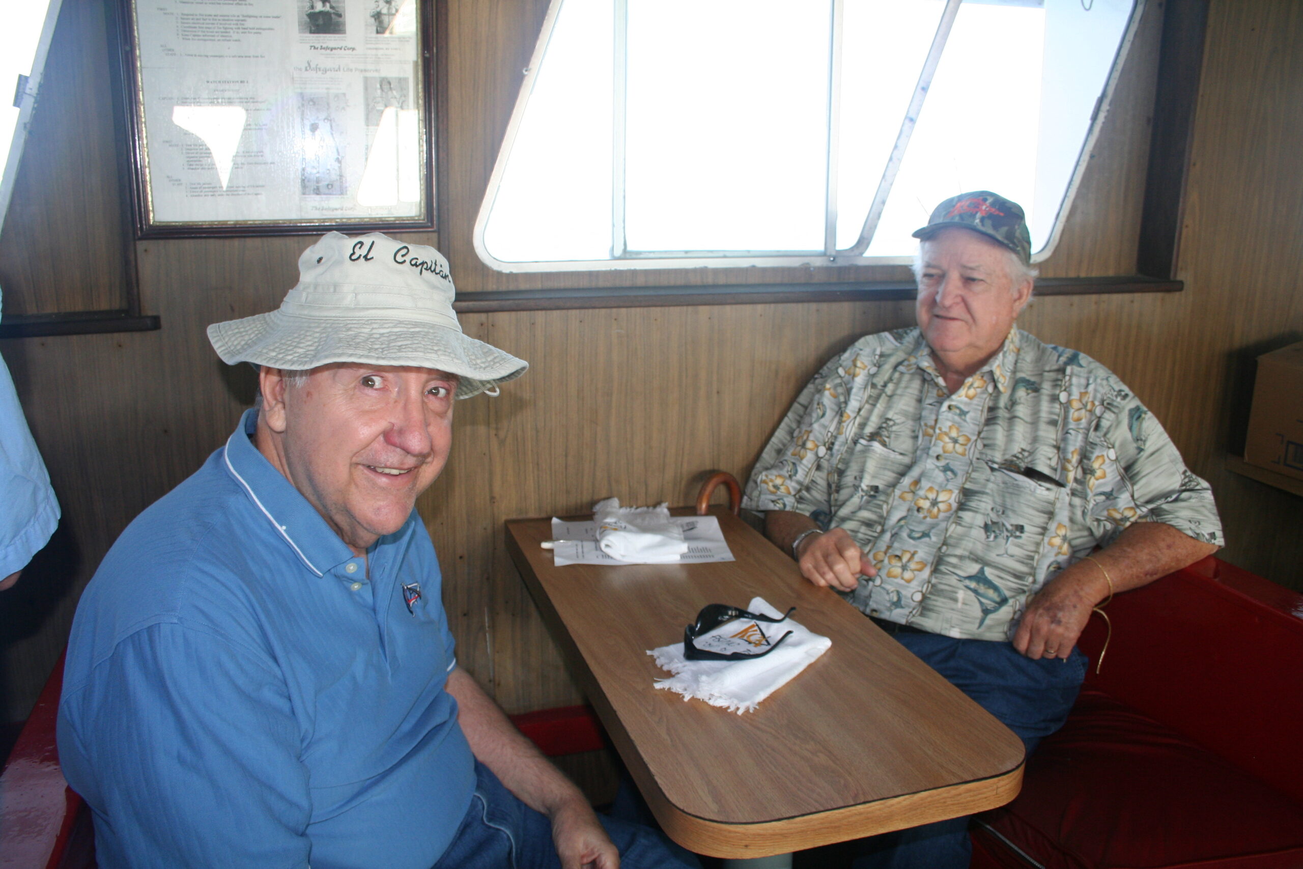 Two men sitting at a table on a boat, one wearing a hat with "El Capitan" embroidered. They are smiling and appear relaxed