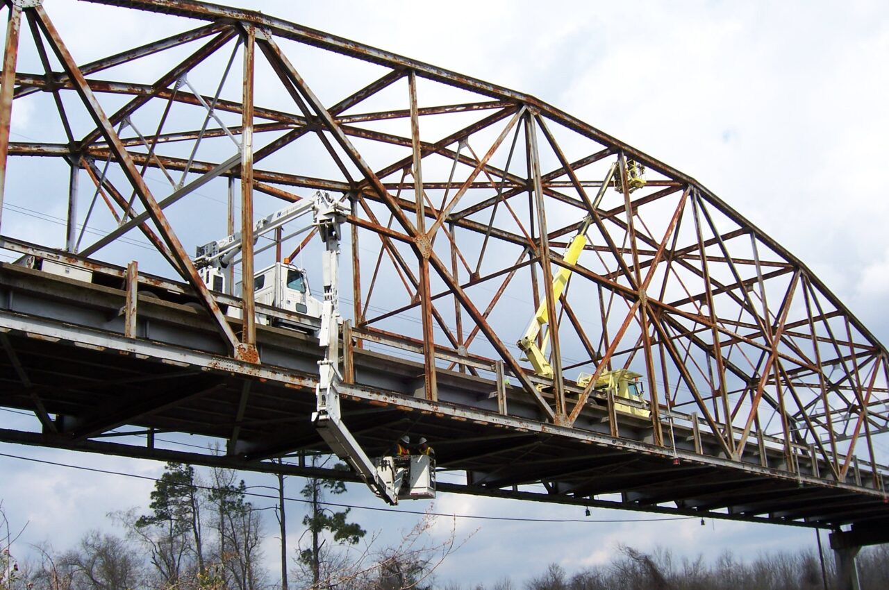 Utility truck inspecting rusty truss bridge over forested area