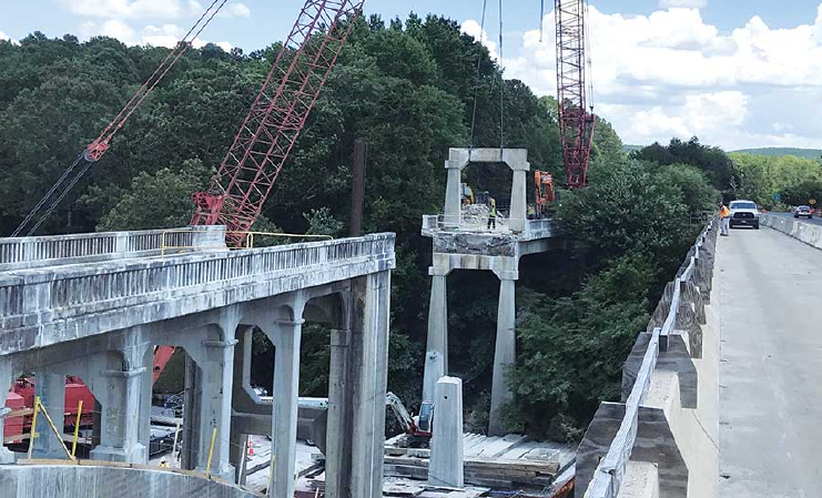 Bridge construction site with cranes lifting concrete structures amidst trees, blue sky, and vehicles passing on adjacent road