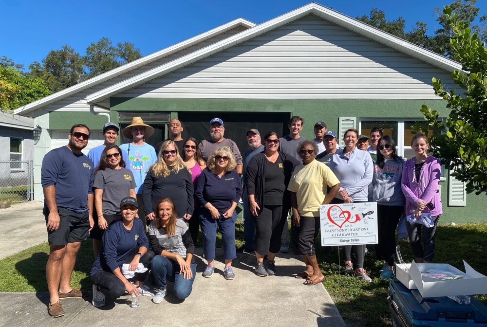 Group of people standing in front of a house, holding a sign: "Paint Your Heart Out Clearwater" with signatures