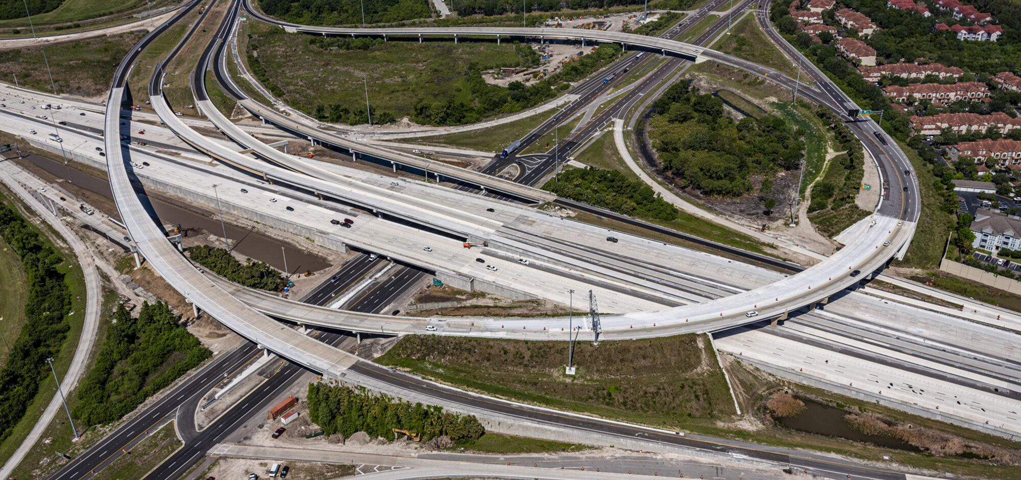 Aerial view of a multi-level highway interchange with surrounding greenery and buildings, showing active traffic flow