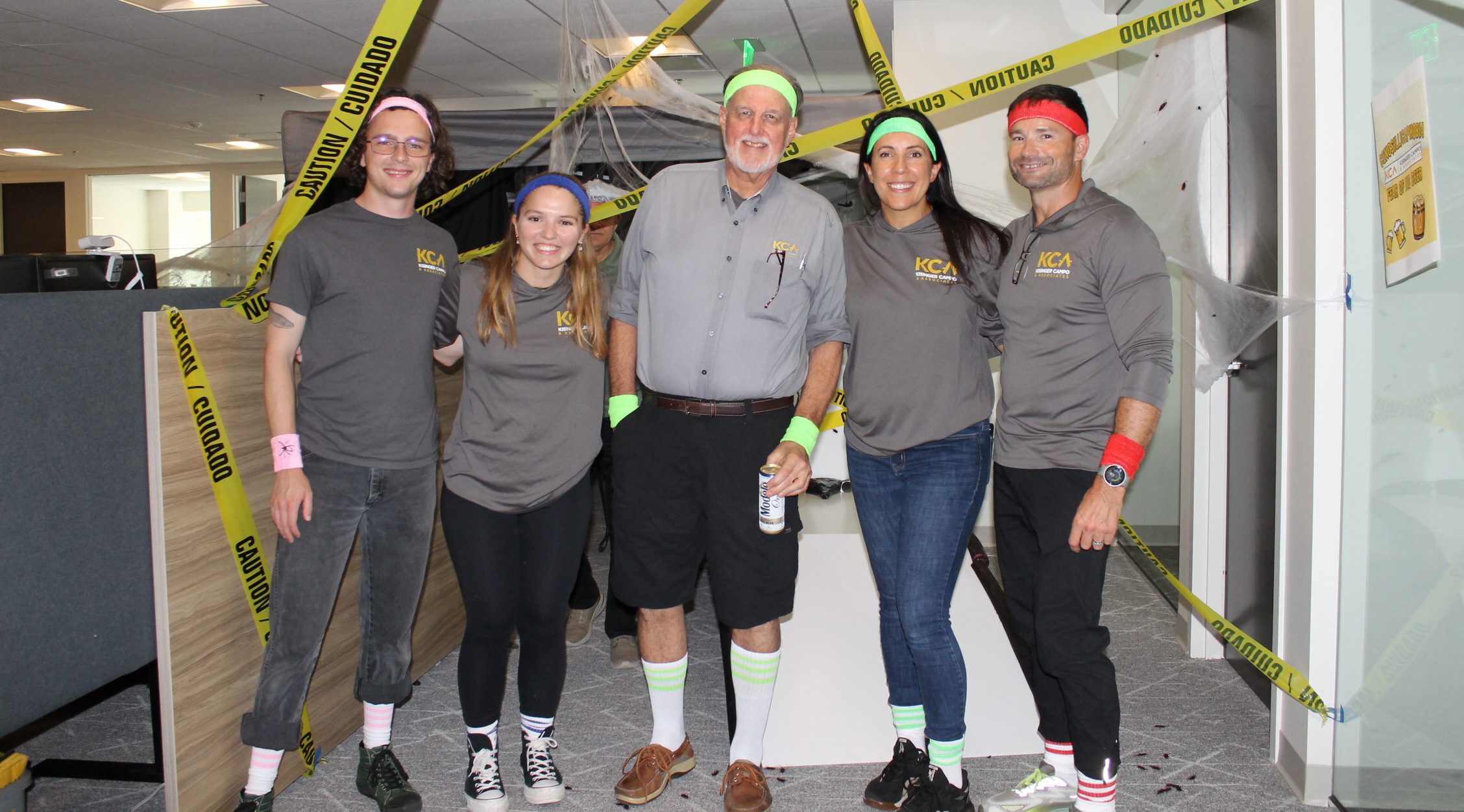 Group of five people in grey KCA shirts with colorful accessories, standing under "Caution" tape in an office setting