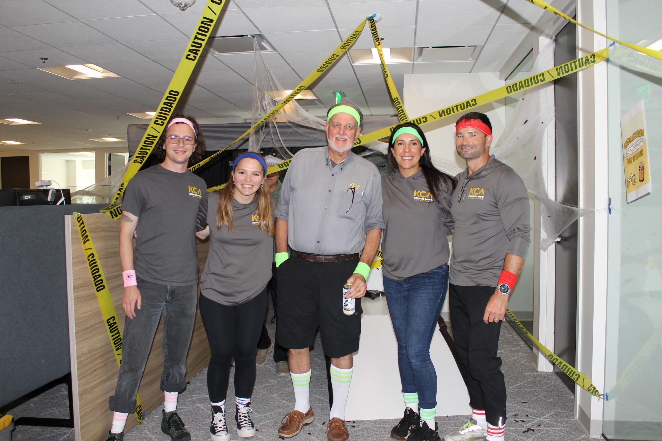 Group of five people in grey KCA shirts with colorful accessories, standing under "Caution" tape in an office setting