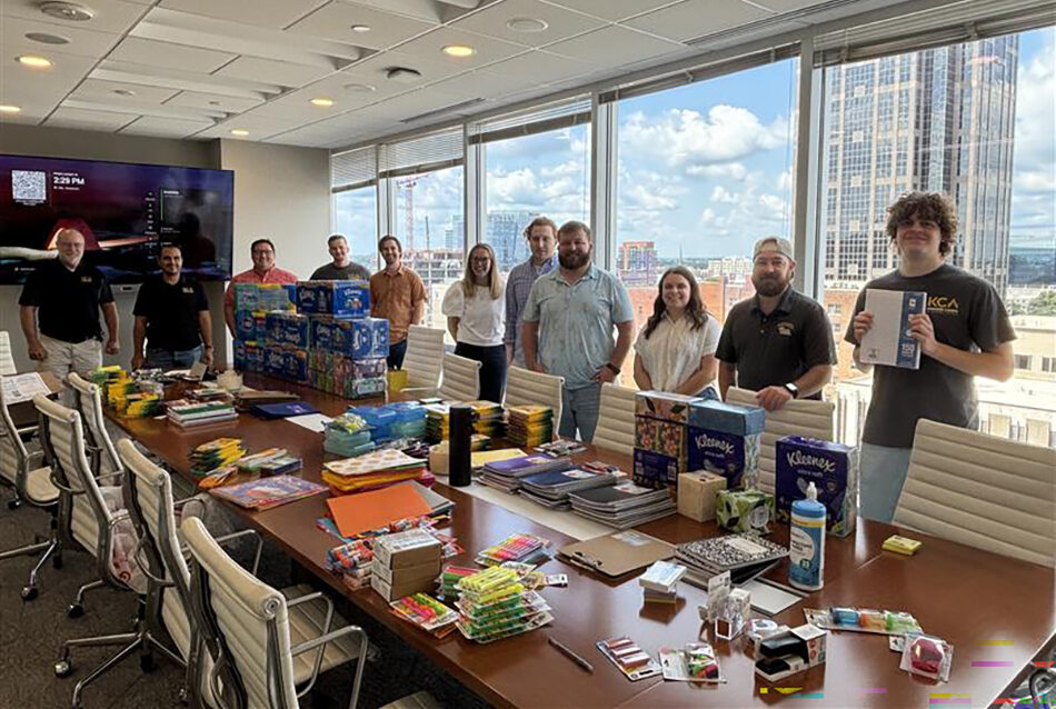 Group of ten people standing behind a conference table filled with school supplies in a bright office with city view