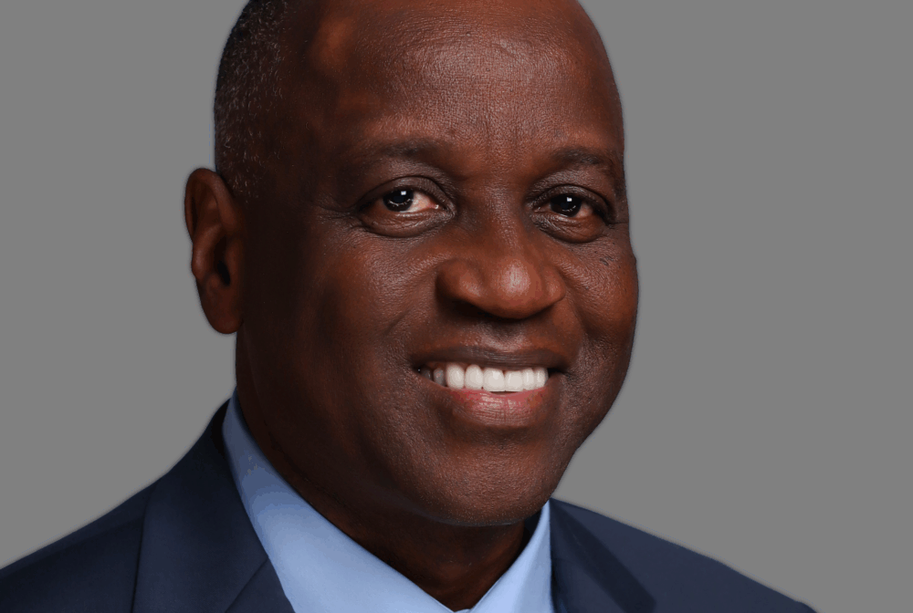 Richard smiling in a blue suit and plaid tie against a gray background