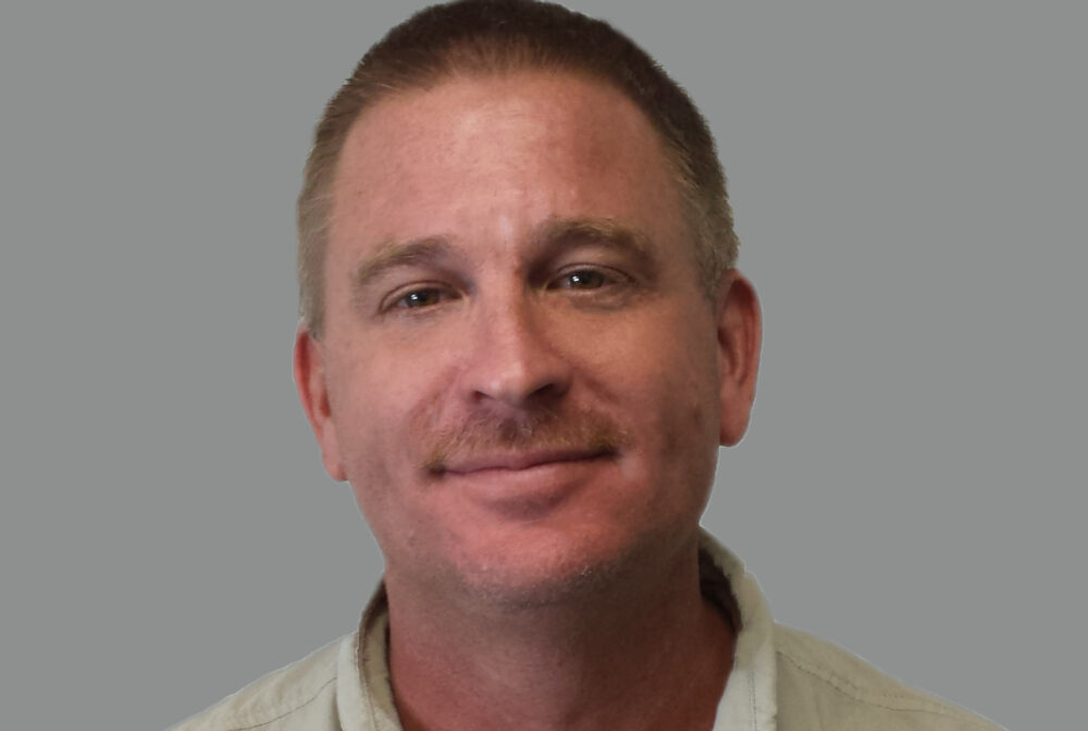 Rye-John.jpg: Smiling man in a light-colored shirt against a gray background