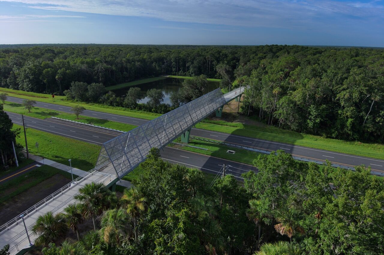 Aerial view of a pedestrian bridge crossing over a road, surrounded by lush greenery and a pond under a blue sky