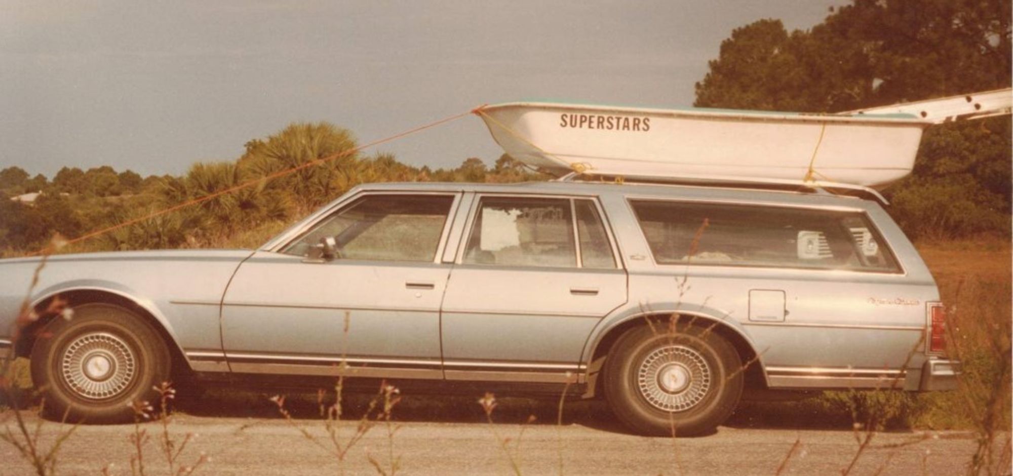 Blue station wagon with a "Superstars" boat on the roof, parked roadside