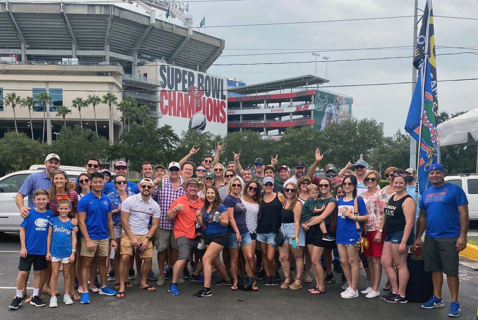 Group of people tailgating outside a stadium with "SUPER BOWL CHAMPIONS" signage. Cornhole game marked "FLORIDA," visible in foreground