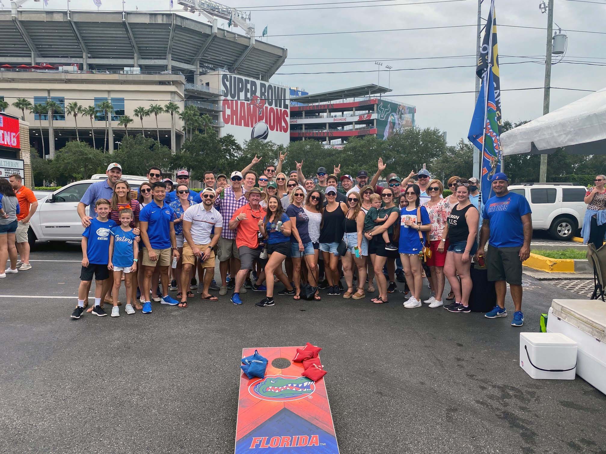 Group of people tailgating outside a stadium with "SUPER BOWL CHAMPIONS" signage. Cornhole game marked "FLORIDA," visible in foreground