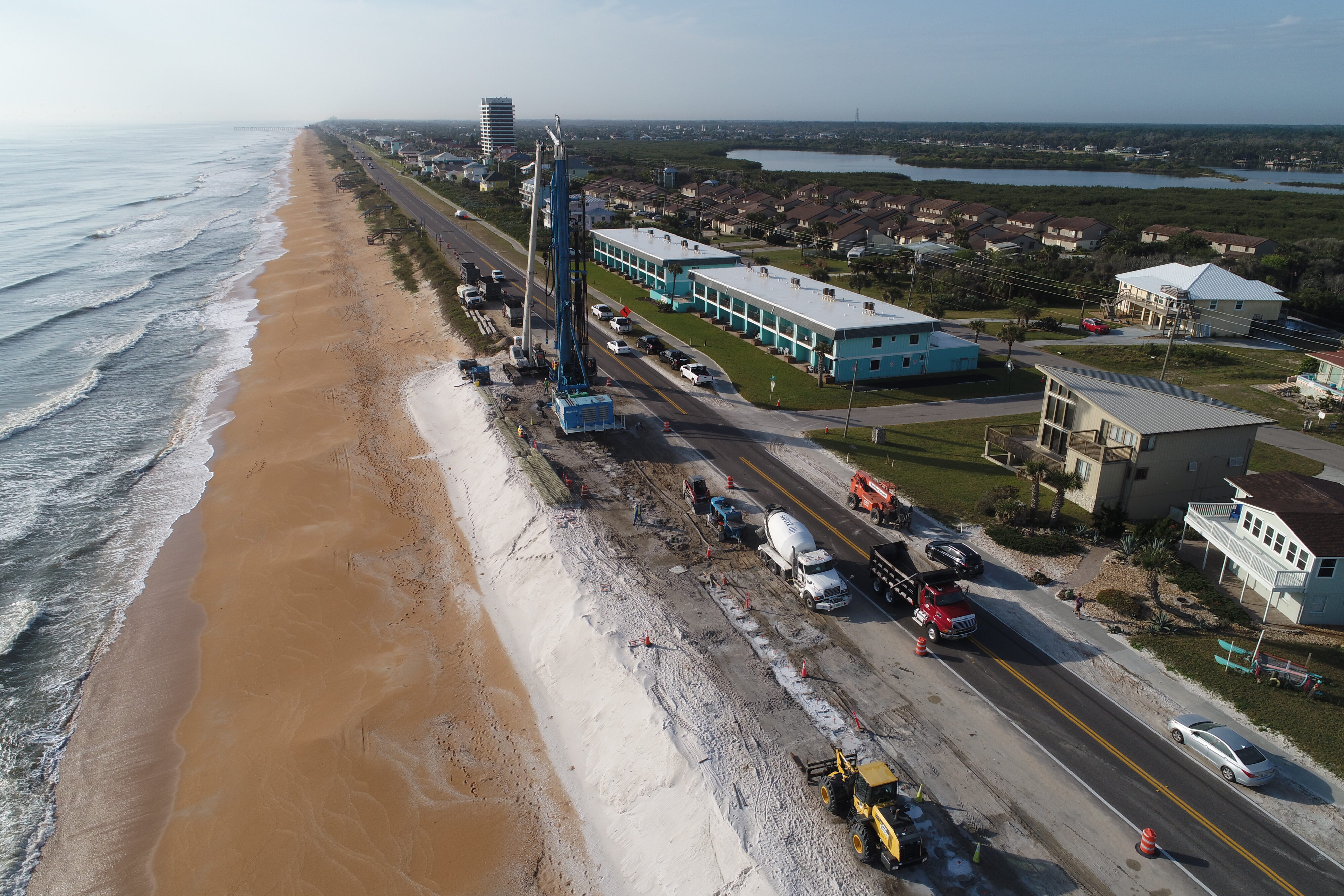 Beachside road construction with vehicles, machinery, and coastal erosion repairs, near residential buildings and a long shoreline