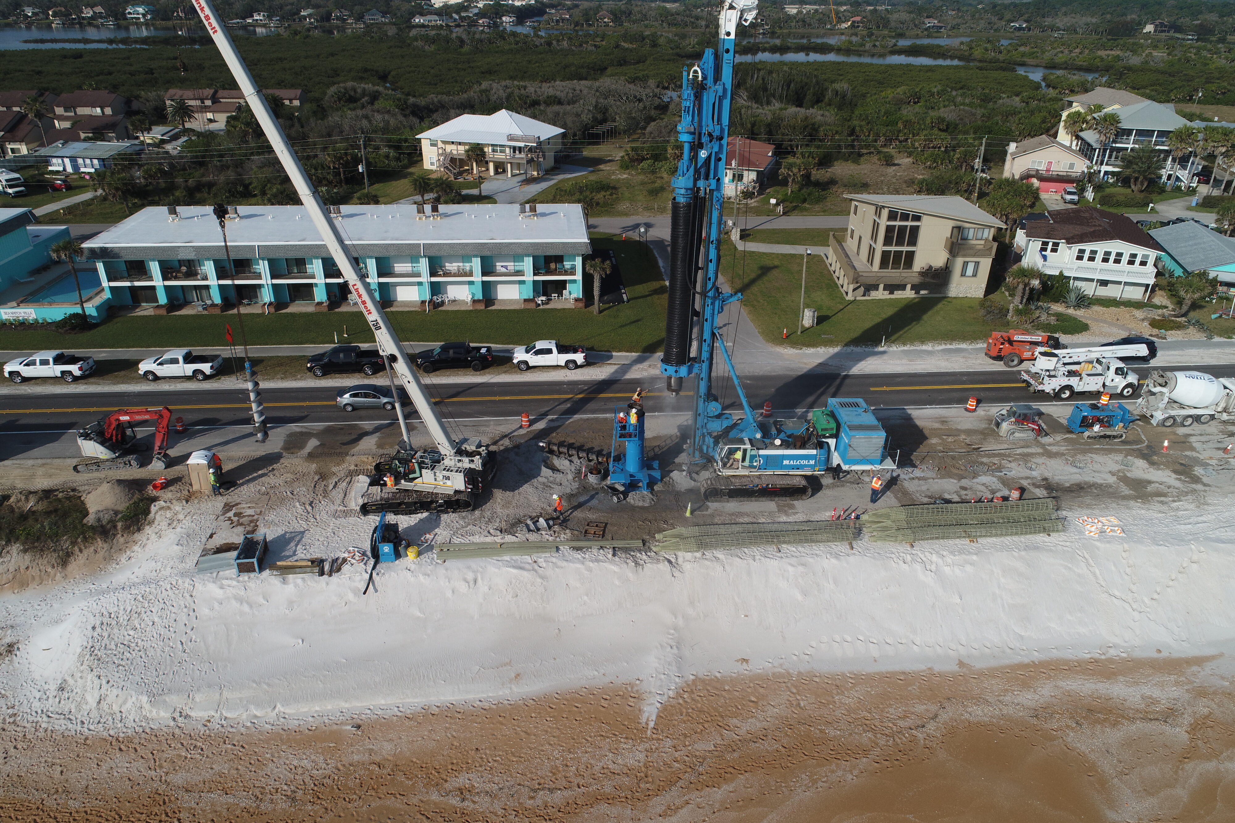 Aerial view of a construction site on a beach. Multiple cranes and vehicles, including a large blue drilling machine, operate alongside a road. Workers in safety gear manage various tasks. Nearby buildings include a turquoise motel and residential homes surrounded by lush greenery