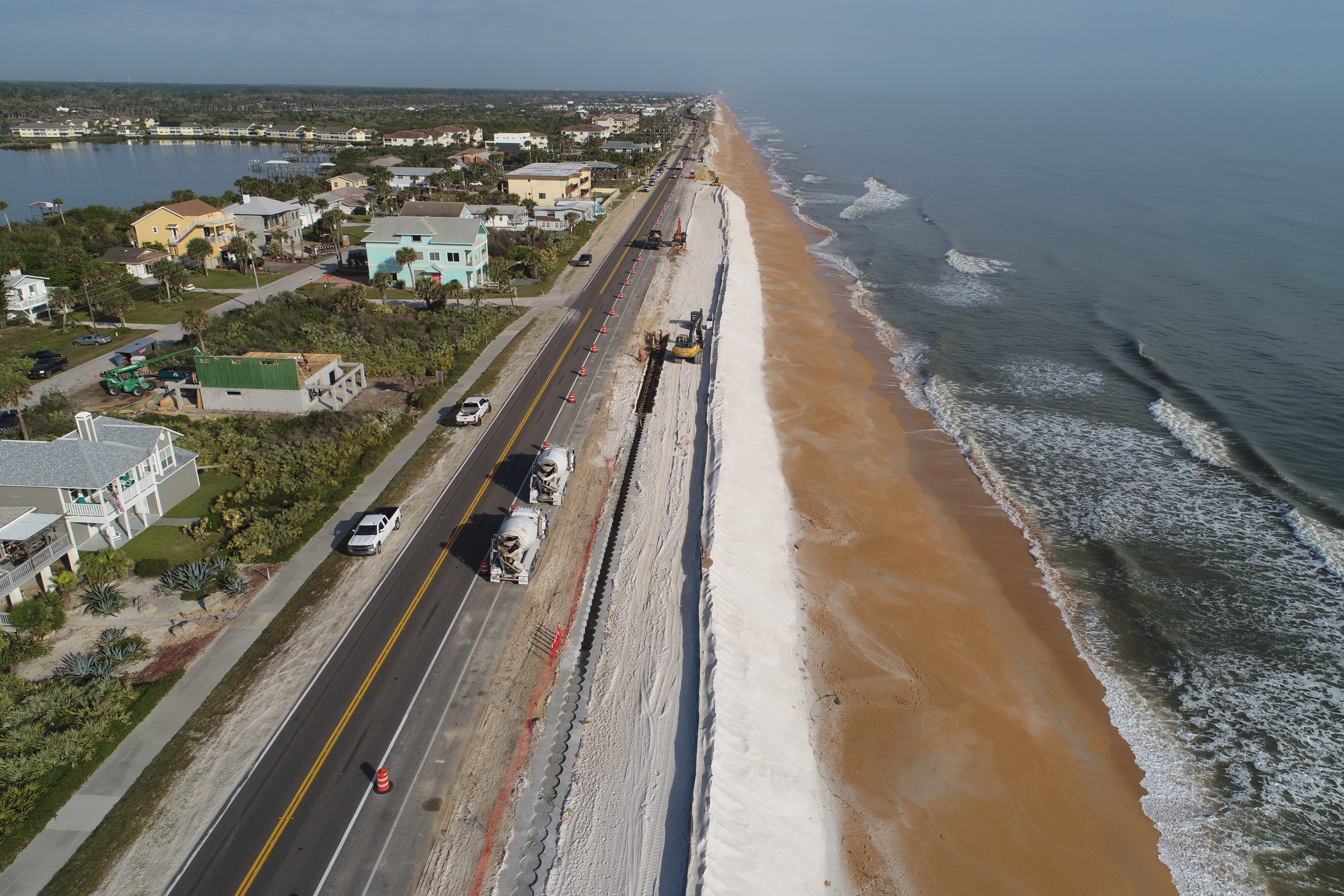 Aerial view of coastline road construction, with machinery and trucks along the beach and residential houses nearby