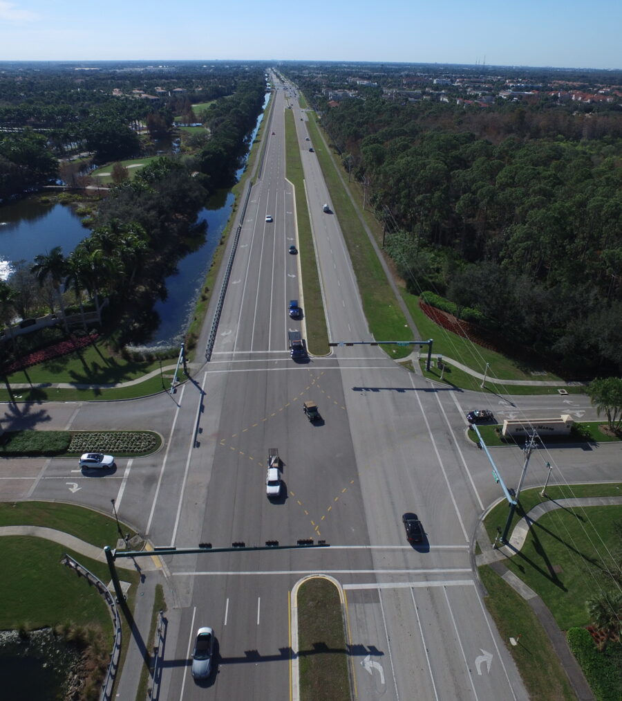 Aerial view of a wide intersection with sparse traffic, surrounded by trees and greenery, under a clear blue sky