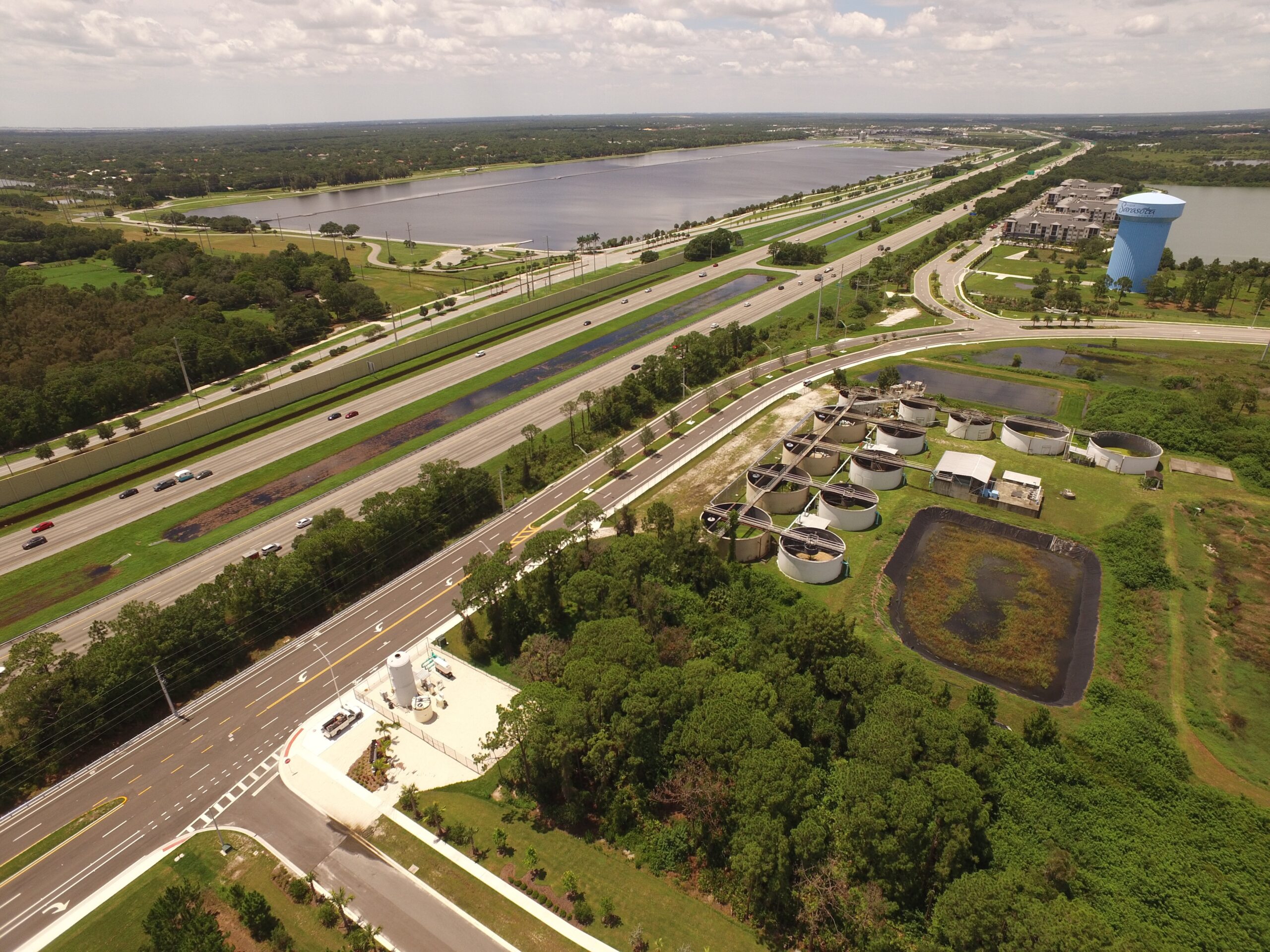 Aerial view of a water treatment plant next to a highway with multiple circular tanks, surrounded by greenery and nearby water bodies