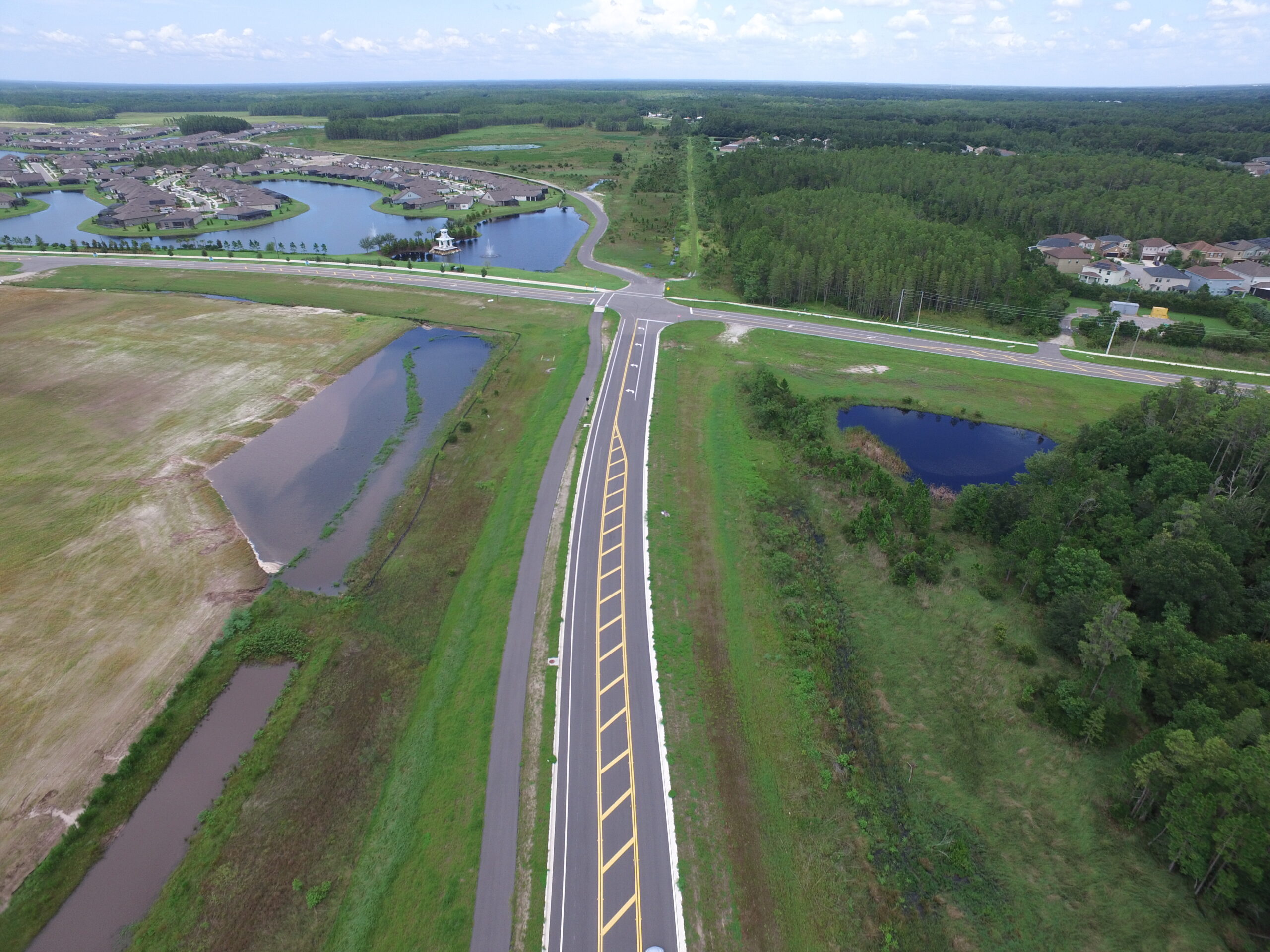 Aerial view of a residential area with surrounding green landscape and ponds, intersected by a wide road