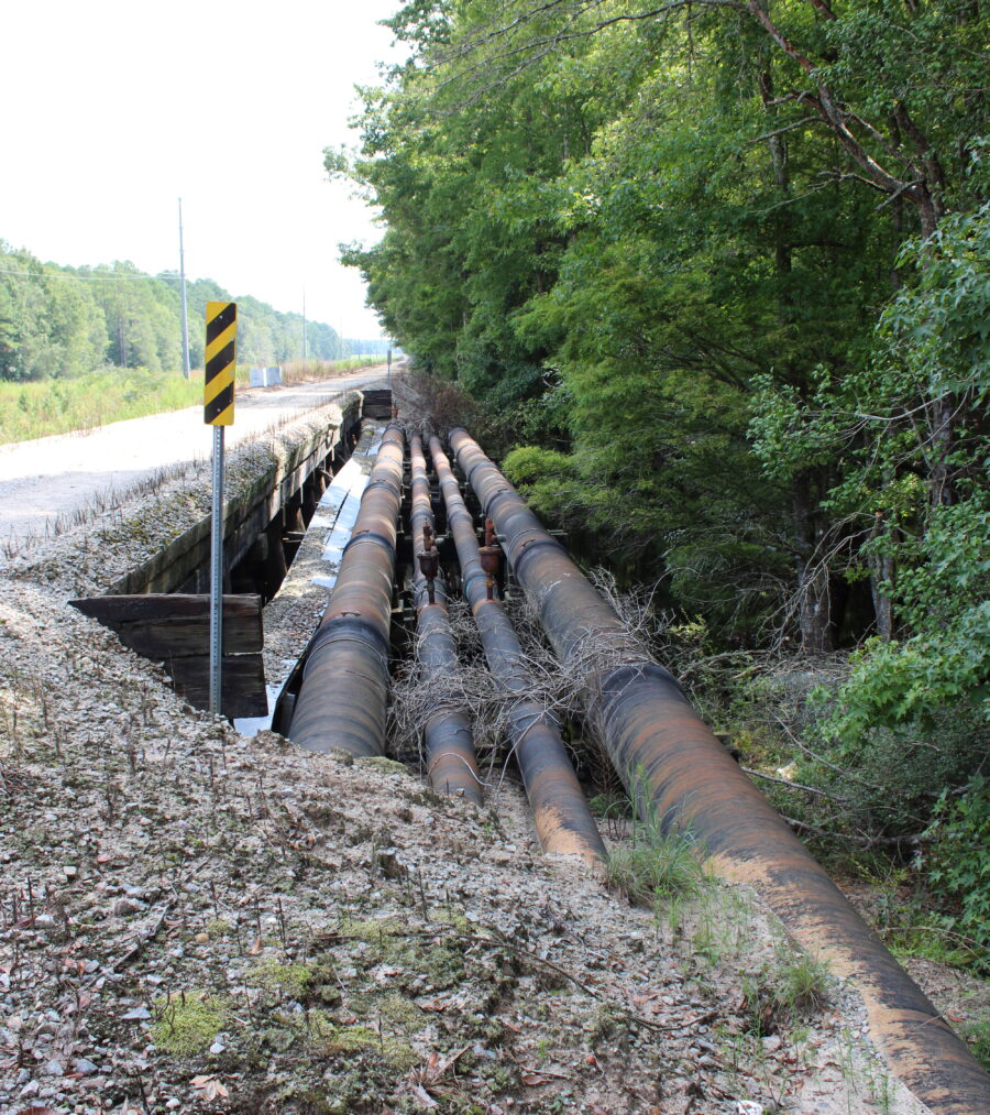 Large rusty pipes run parallel to a rural road, bordered by trees