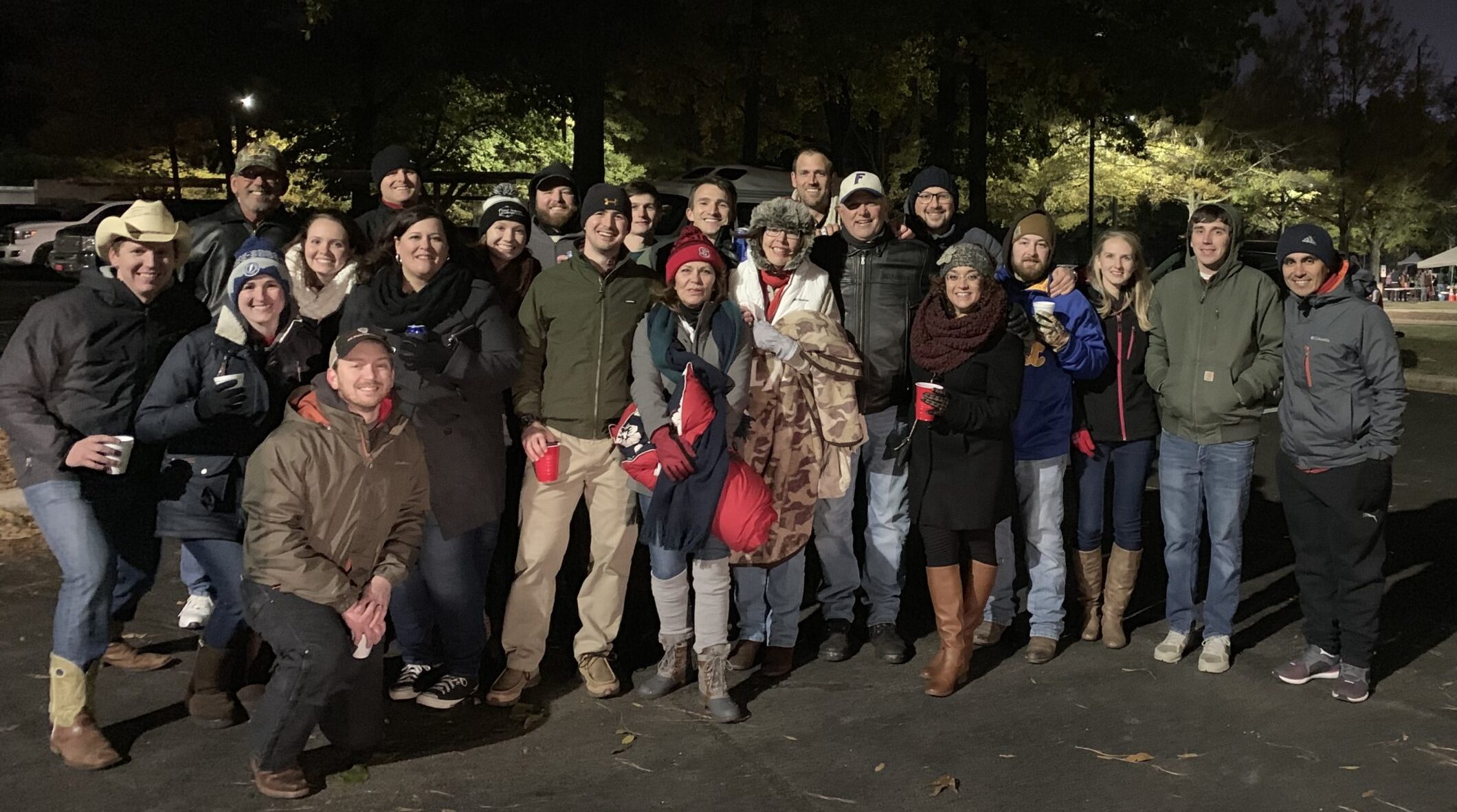 Large group of people gathered outdoors at night, dressed warmly and smiling, with a dark tree-lined background