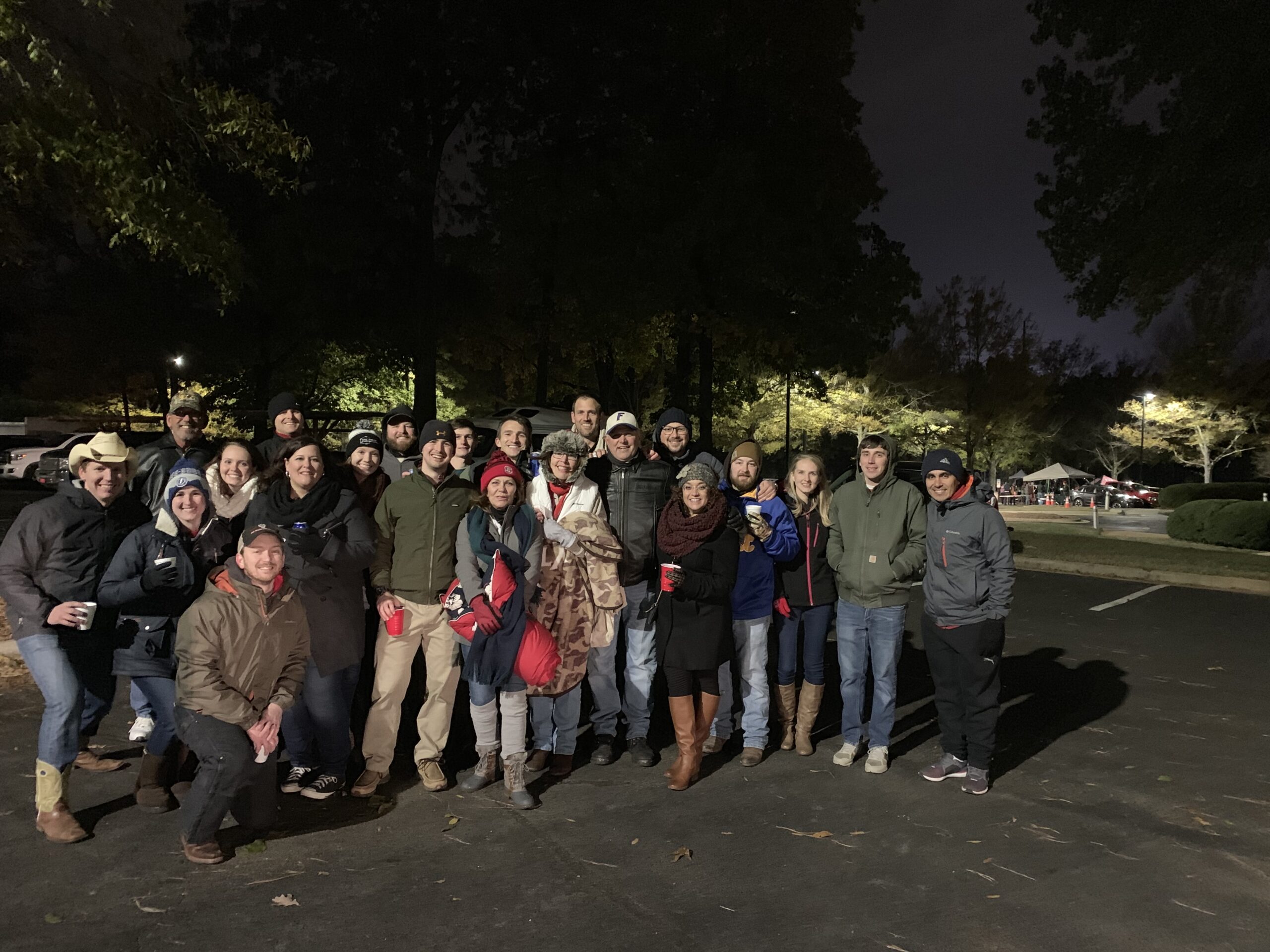 Large group of people gathered outdoors at night, dressed warmly and smiling, with a dark tree-lined background