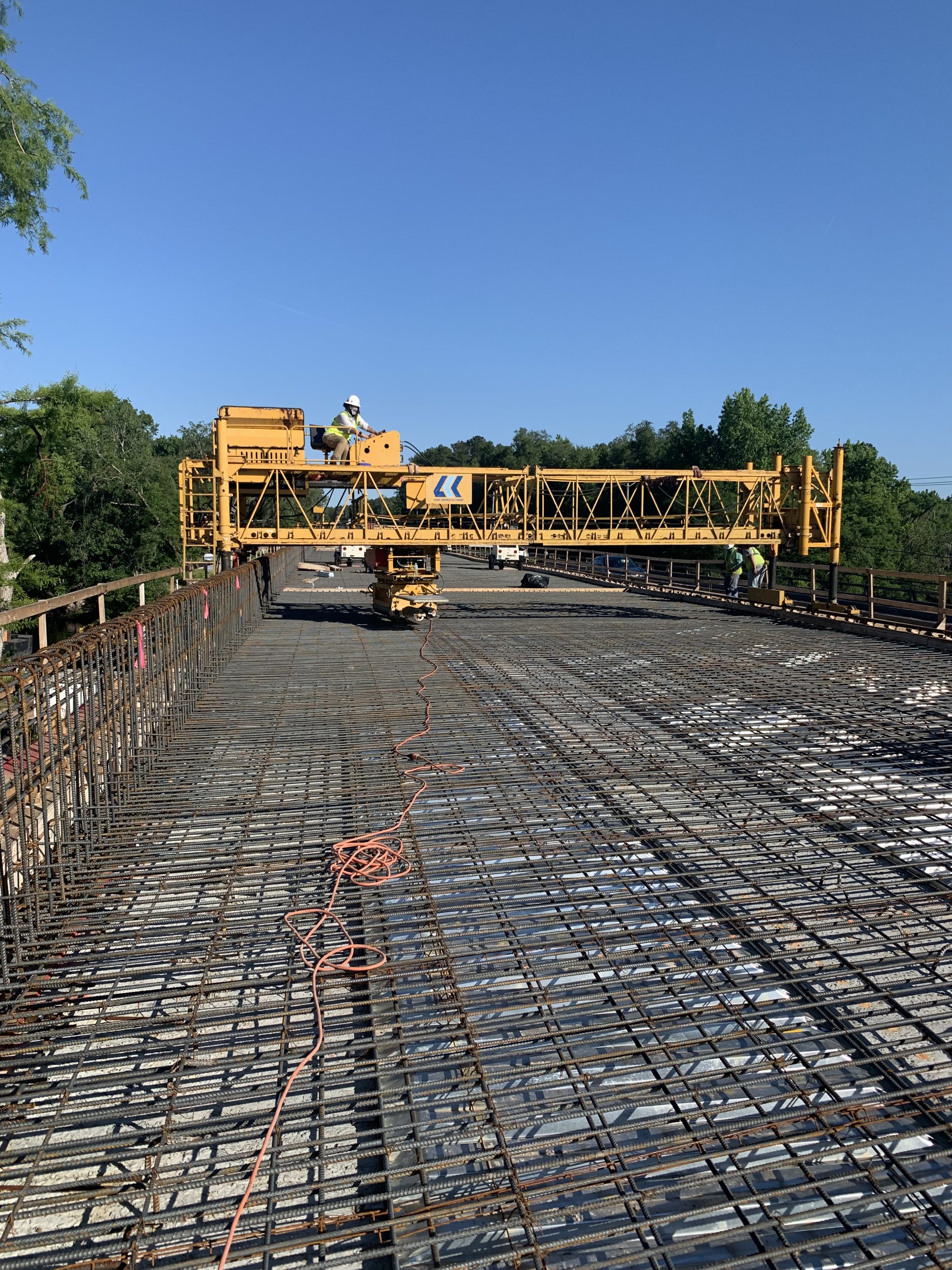 Construction workers on a bridge, operating a large yellow crane with metal beams and blue sky