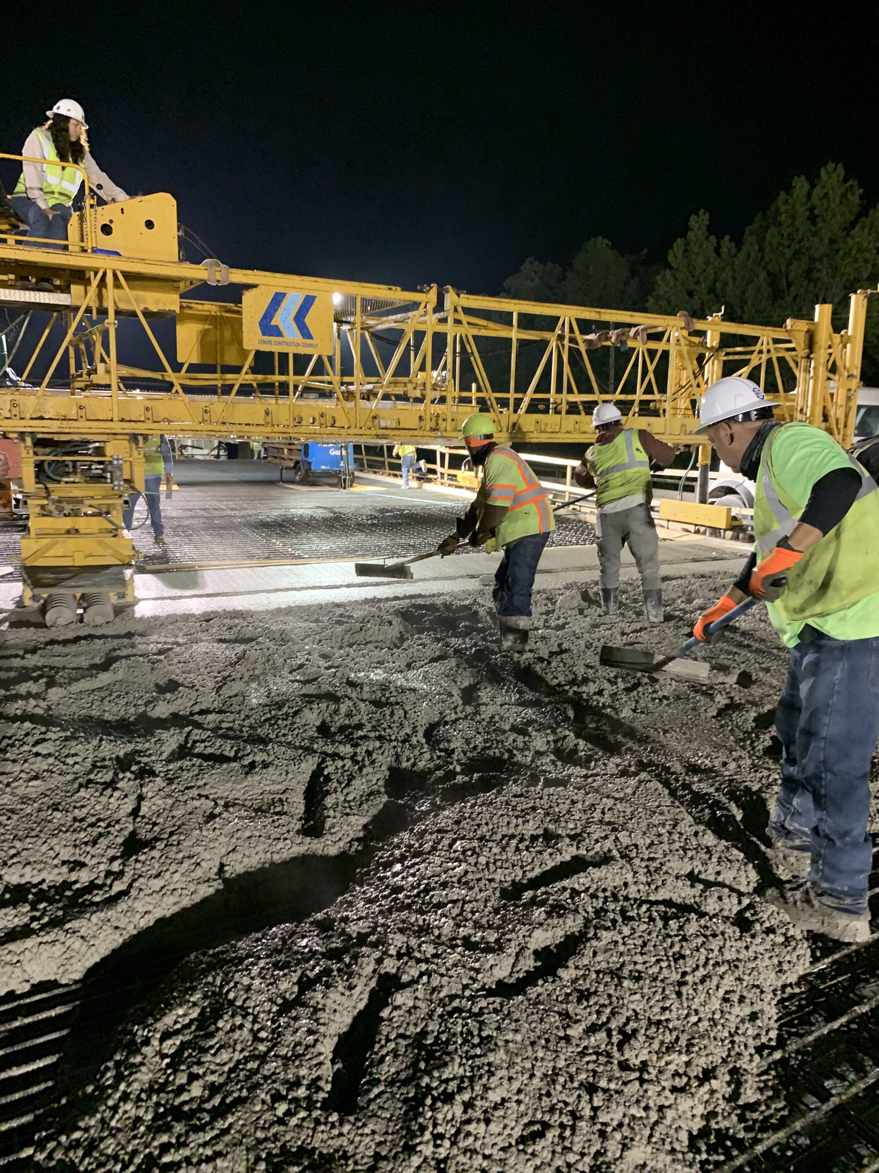 Workers smoothing wet concrete at night under construction equipment
