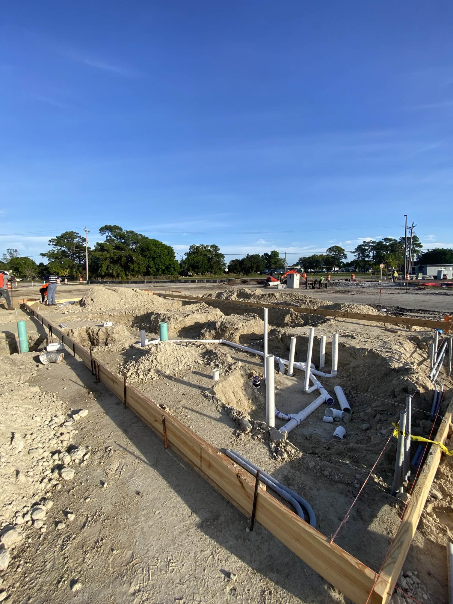 Construction site with piping preparations, wooden framing, and workers in the background under clear blue sky