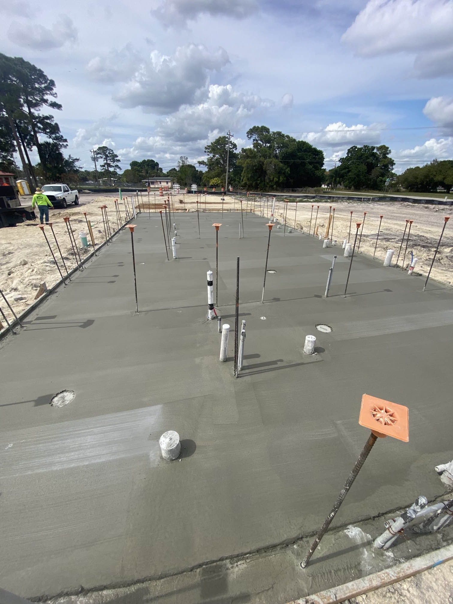 Freshly poured concrete foundation with metal rods, pipes protruding, and a worker in the background on a construction site