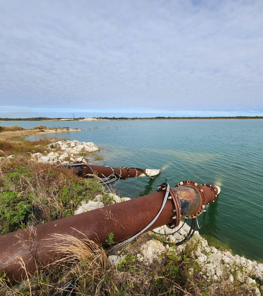 Rusty industrial pipes discharging water into a serene reservoir under a cloudy sky