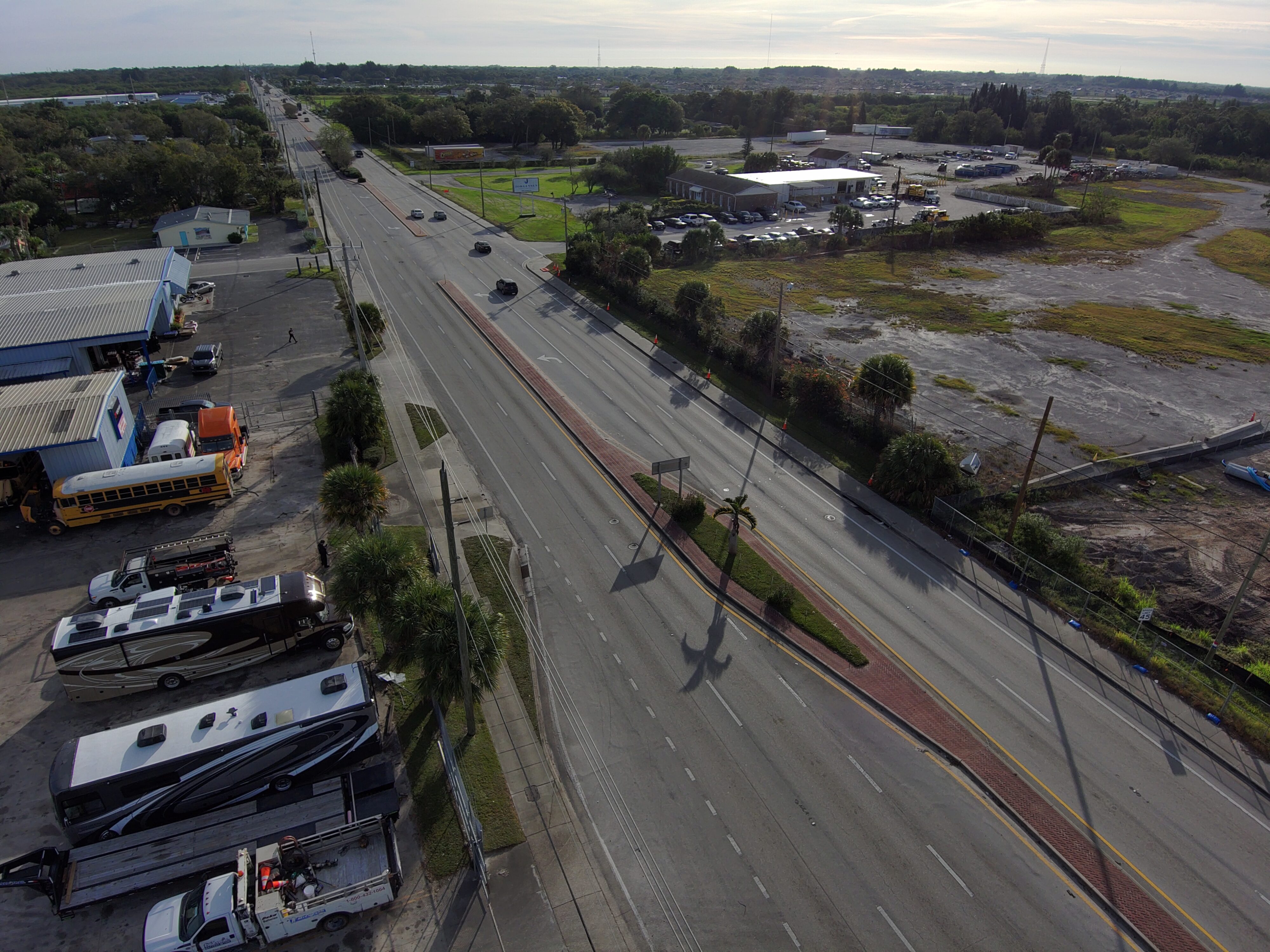 Wide road with sparse traffic, flanked by industrial buildings, parked RVs, and grassy vacant lots