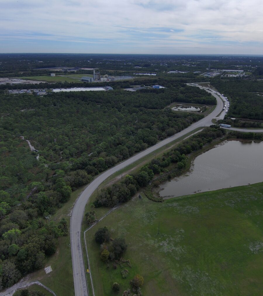 Aerial view of a winding road through dense forest with adjacent water and grassy areas, leading to distant industrial buildings under a cloudy sky