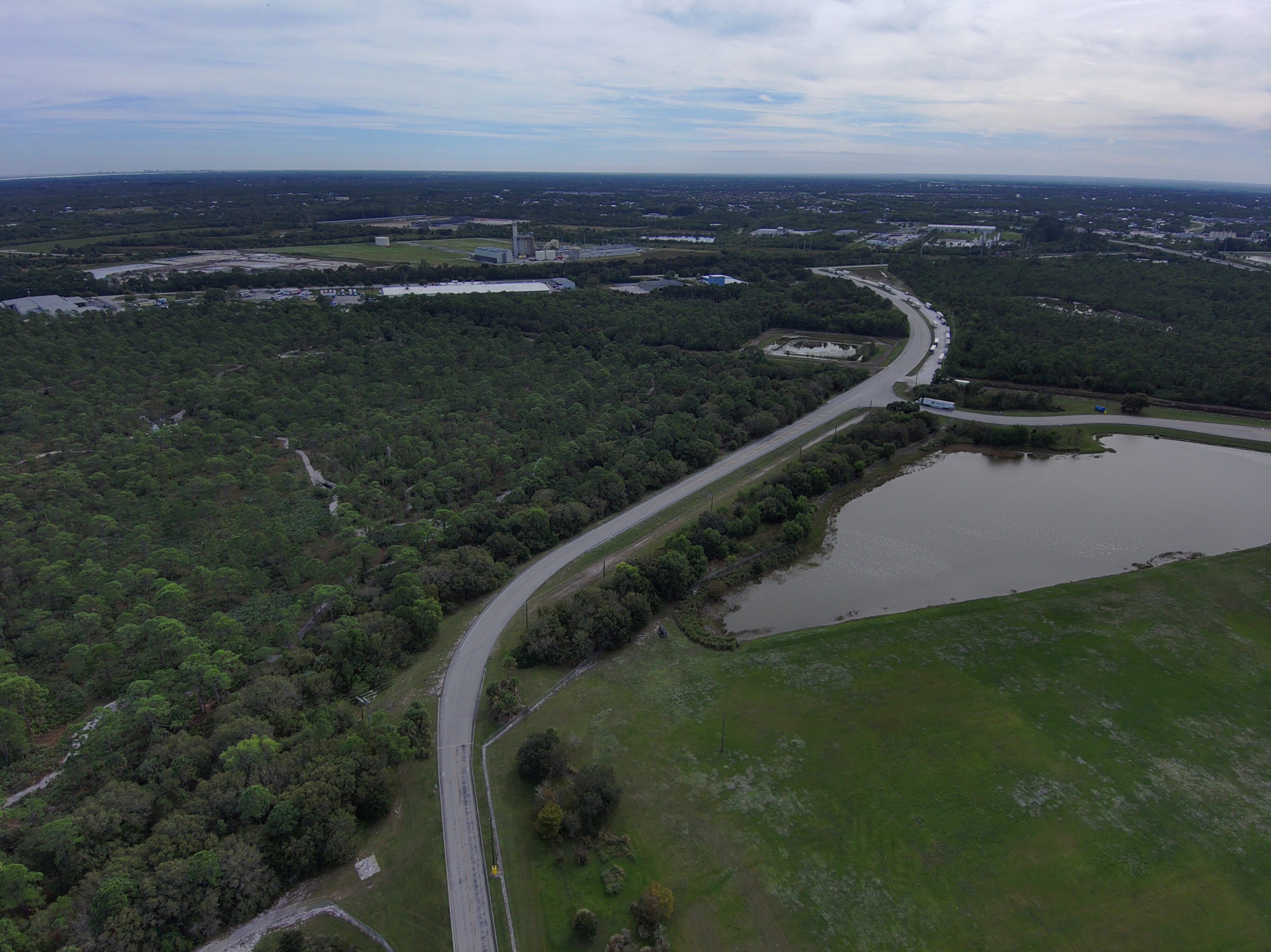 Aerial view of a curving road alongside a forest and lake, with distant industrial buildings in the background