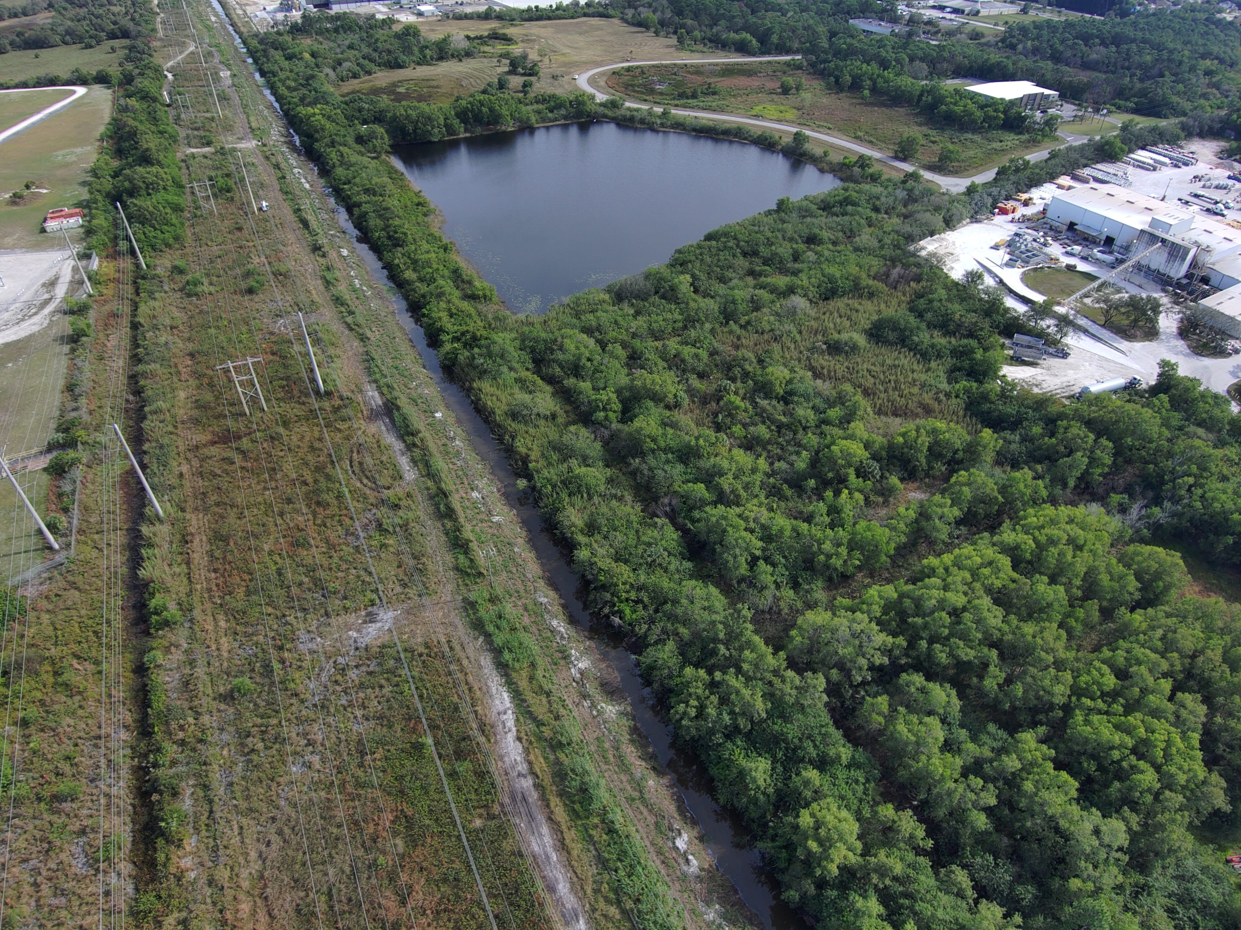Aerial view of power lines beside dense forest, a large pond, and industrial buildings