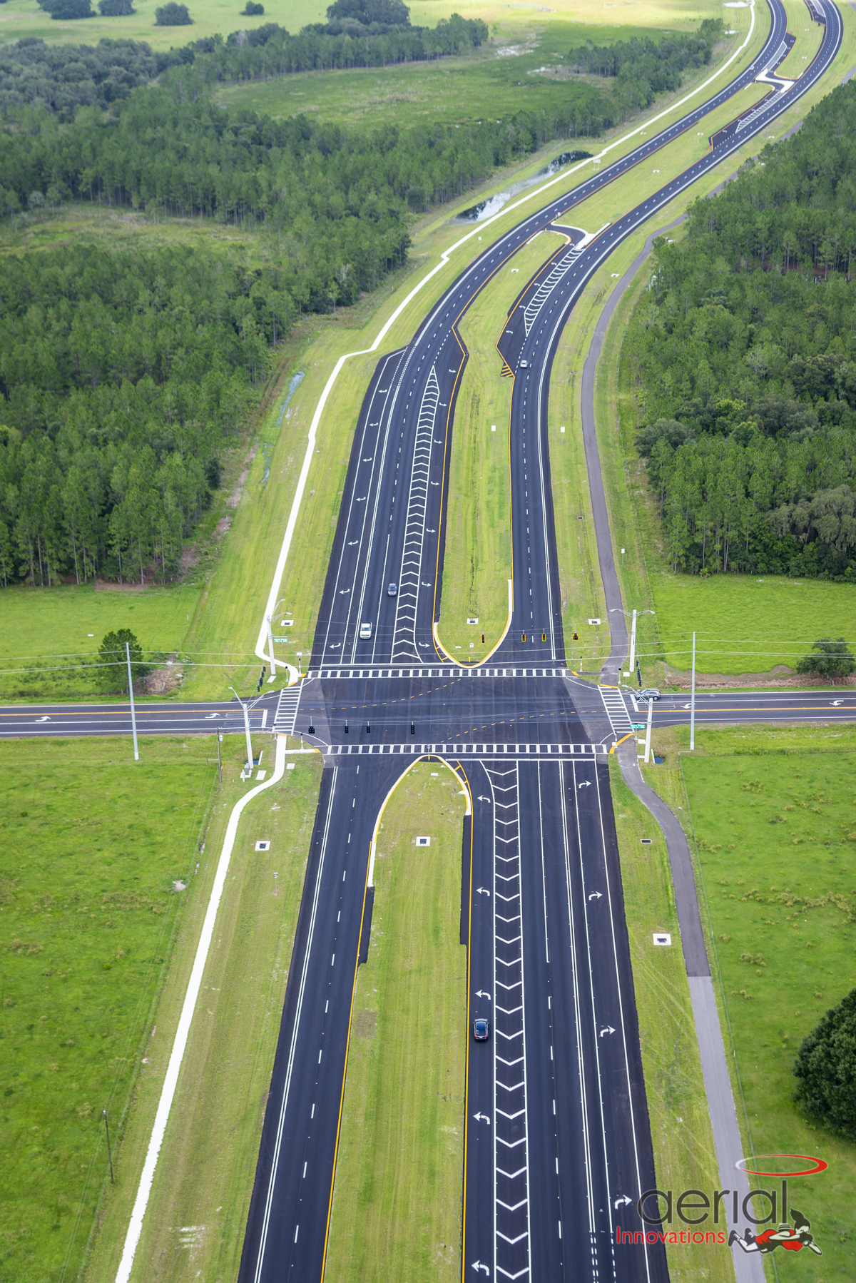 Aerial view of multi-lane divided highway intersection surrounded by green fields, featuring clearly marked lanes and a few vehicles