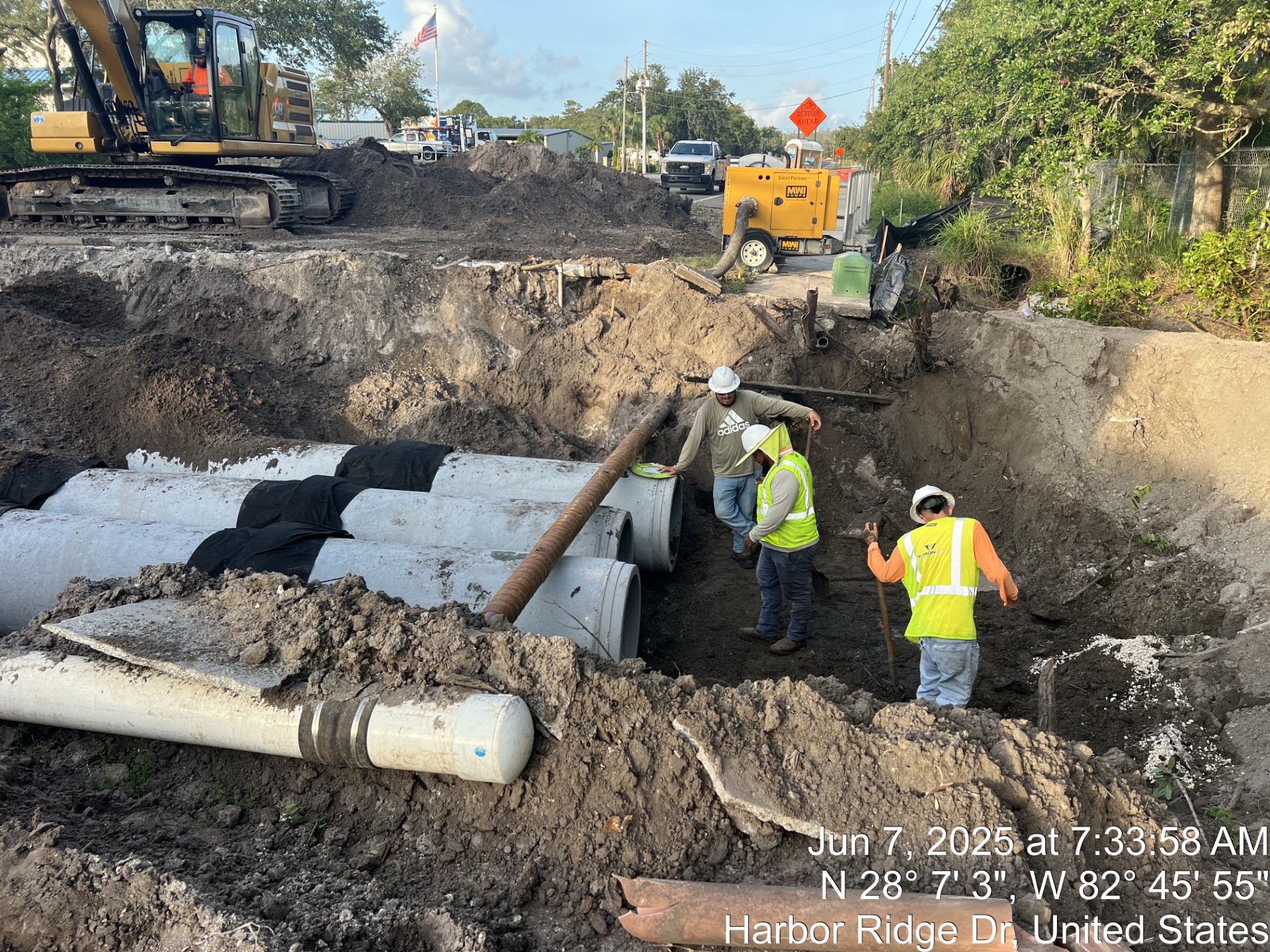 Construction workers install large pipes in an excavated trench, with machinery in the background