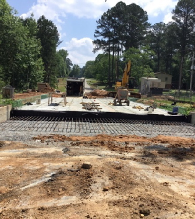 Construction site with heavy machinery, concrete foundation, surrounded by trees and dirt