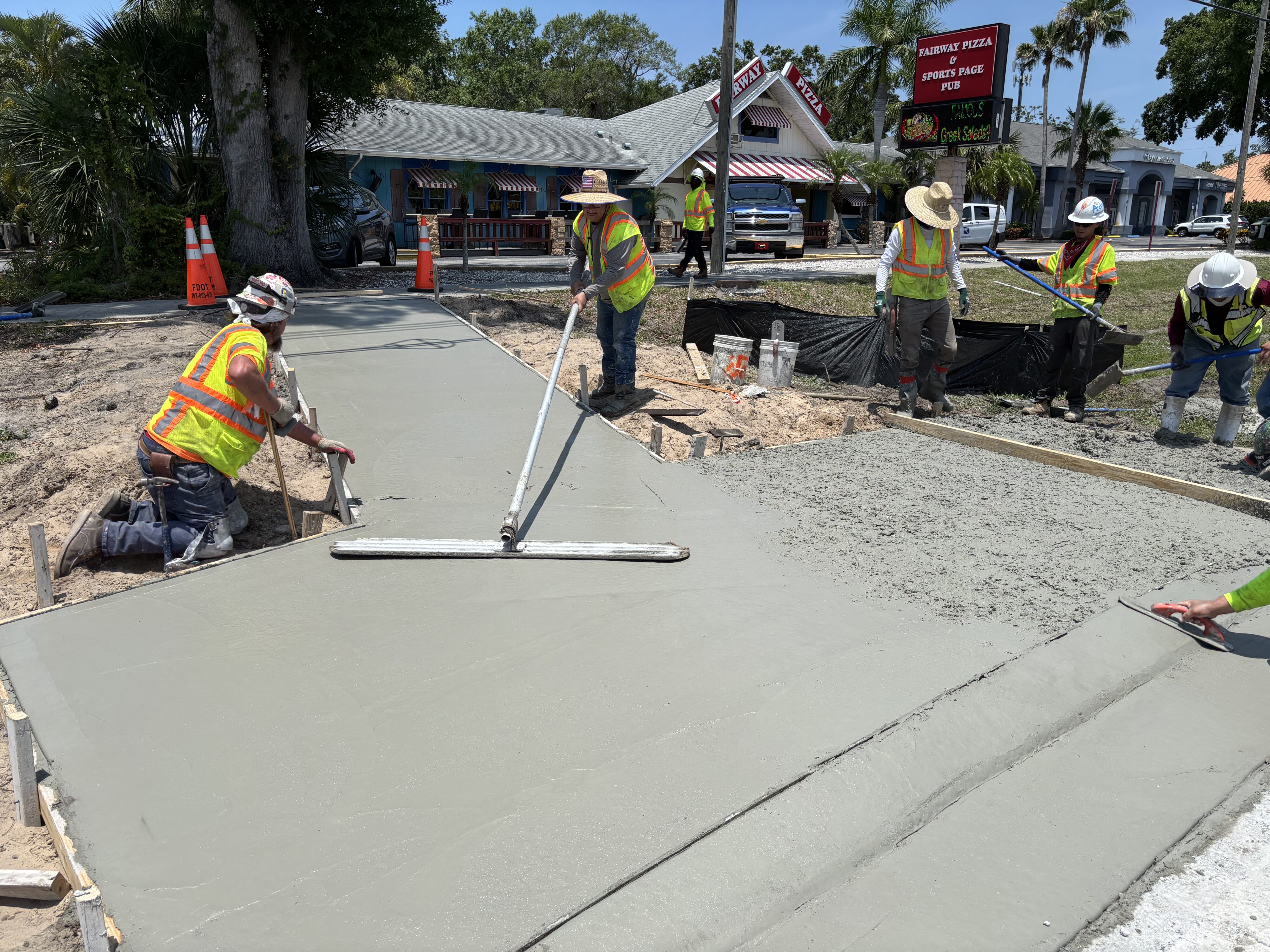 Construction workers smoothing freshly poured concrete on a sidewalk; Fairway Pizza sign in background