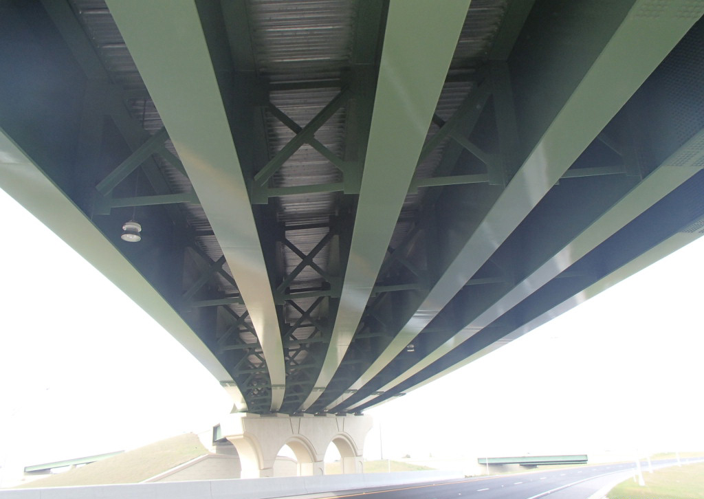 Underside view of a large steel bridge, showcasing green beams and cross-braces, above a highway