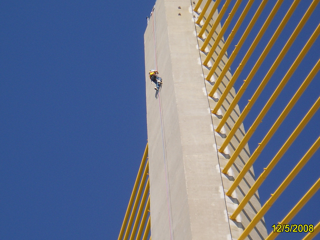 Person climbing a tall structure with striking yellow cables against a clear blue sky