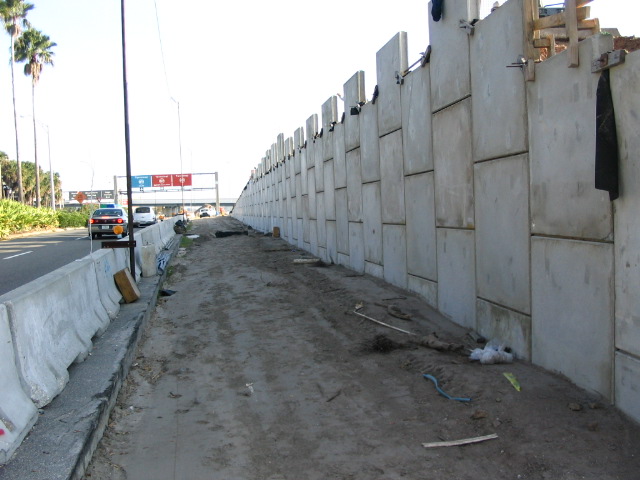 Concrete retaining wall under construction alongside a highway, traffic visible in the distance