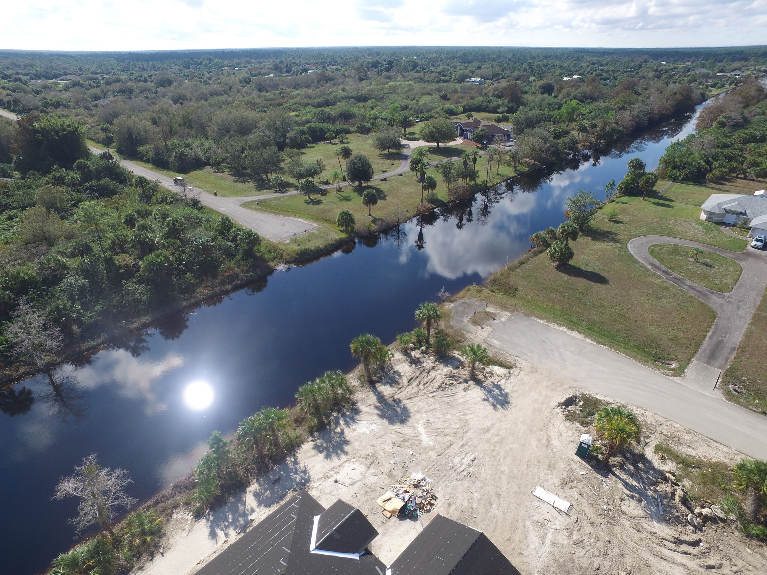 Aerial view of a serene canal reflecting the sky, surrounded by lush greenery and roads in a suburban area