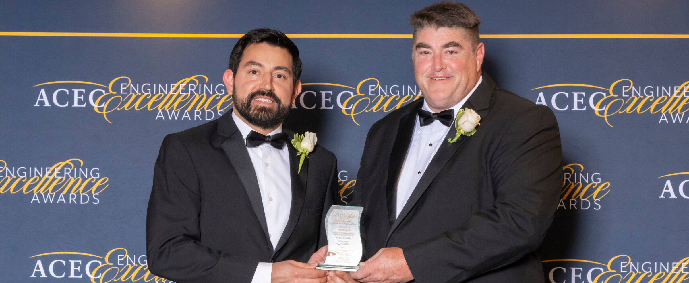 Two men in tuxedos holding an award at ACEC Engineering Excellence Awards event backdrop