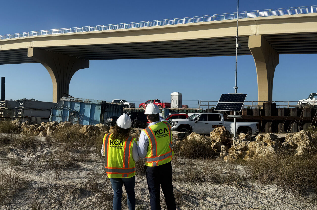 Two people in construction gear with "KCA" vests, observing a bridge construction site with vehicles and equipment