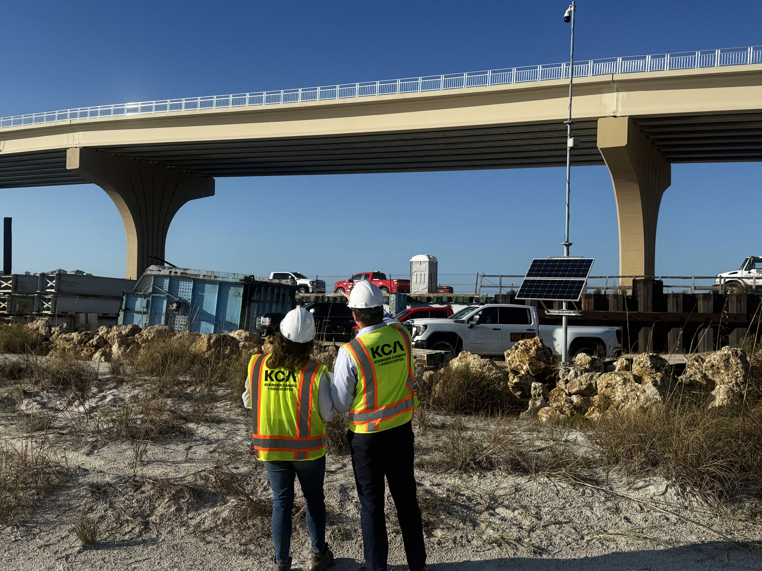 Two people in construction gear with "KCA" vests, observing a bridge construction site with vehicles and equipment