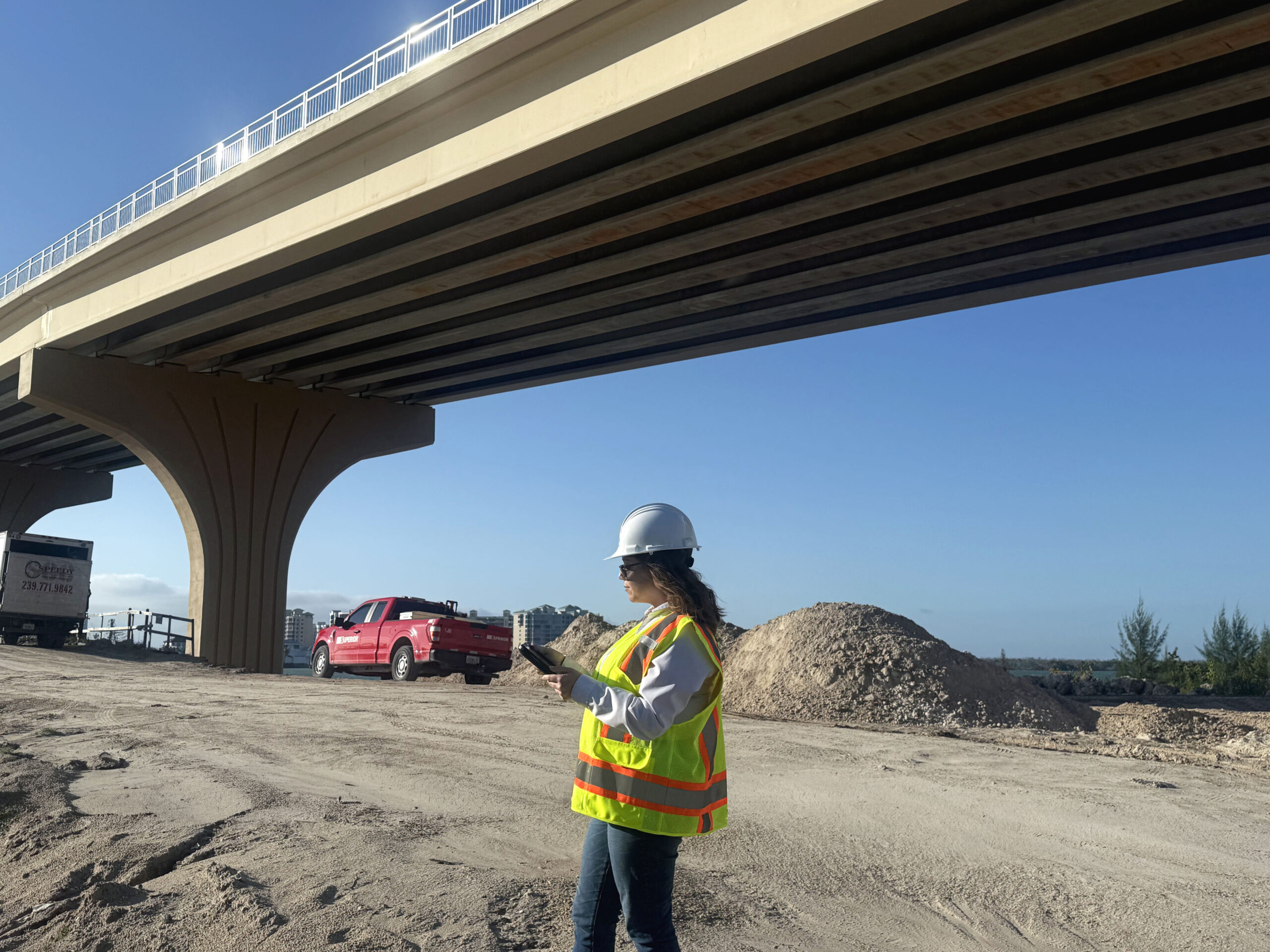 Big Carlos inspects construction site under bridge, wearing safety gear, holding a tablet, with a red truck nearby