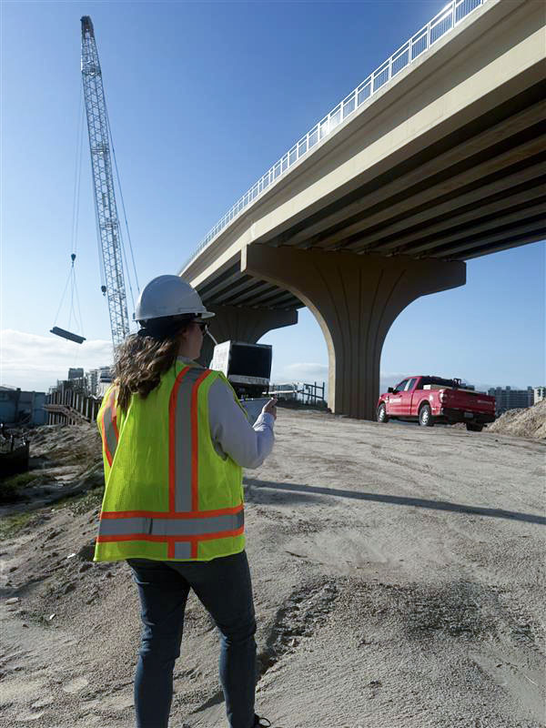 Person in safety gear inspecting bridge construction site with crane and red pickup truck nearby